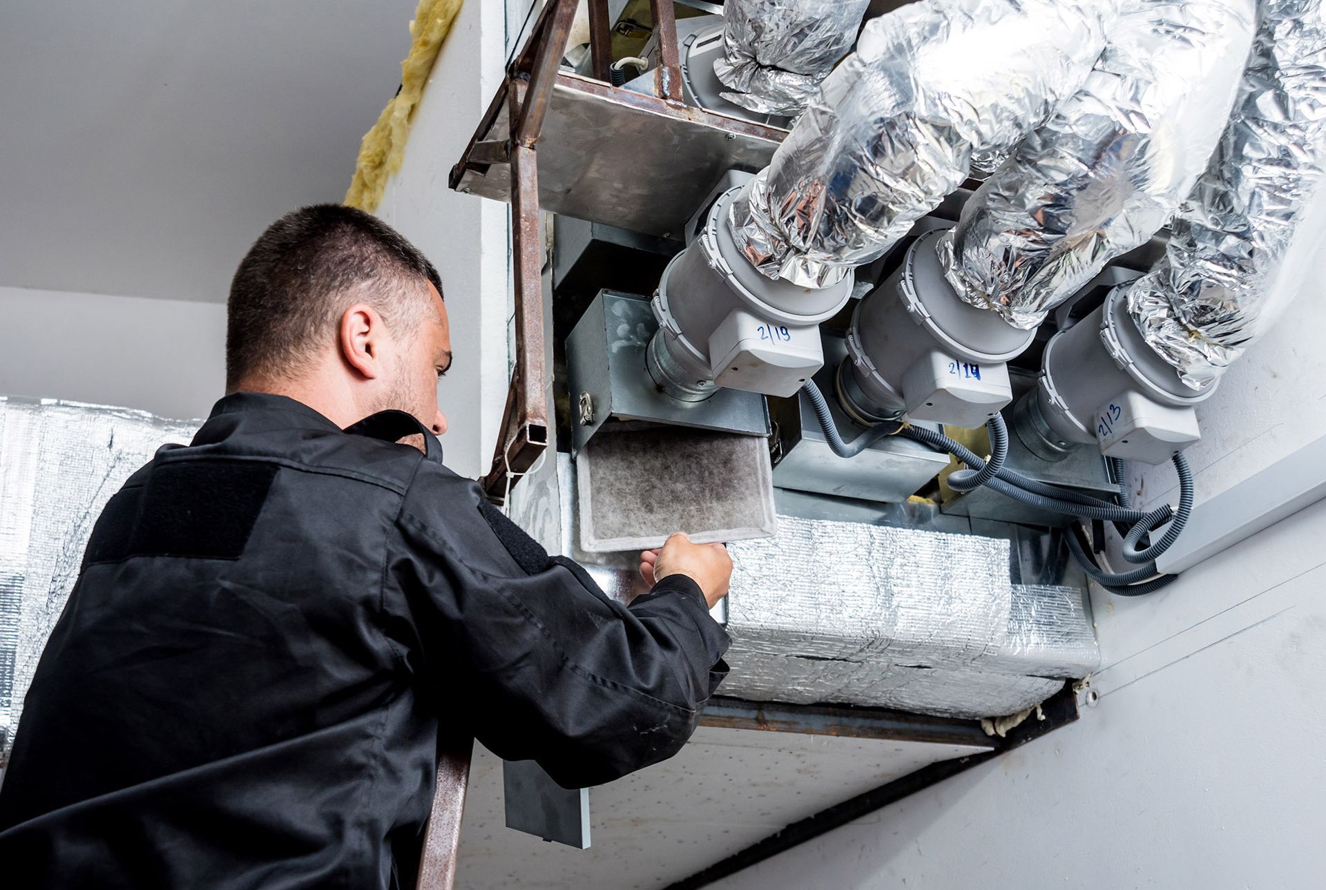 HVAC technician in black uniform working on ventilation system with insulated ducts.
