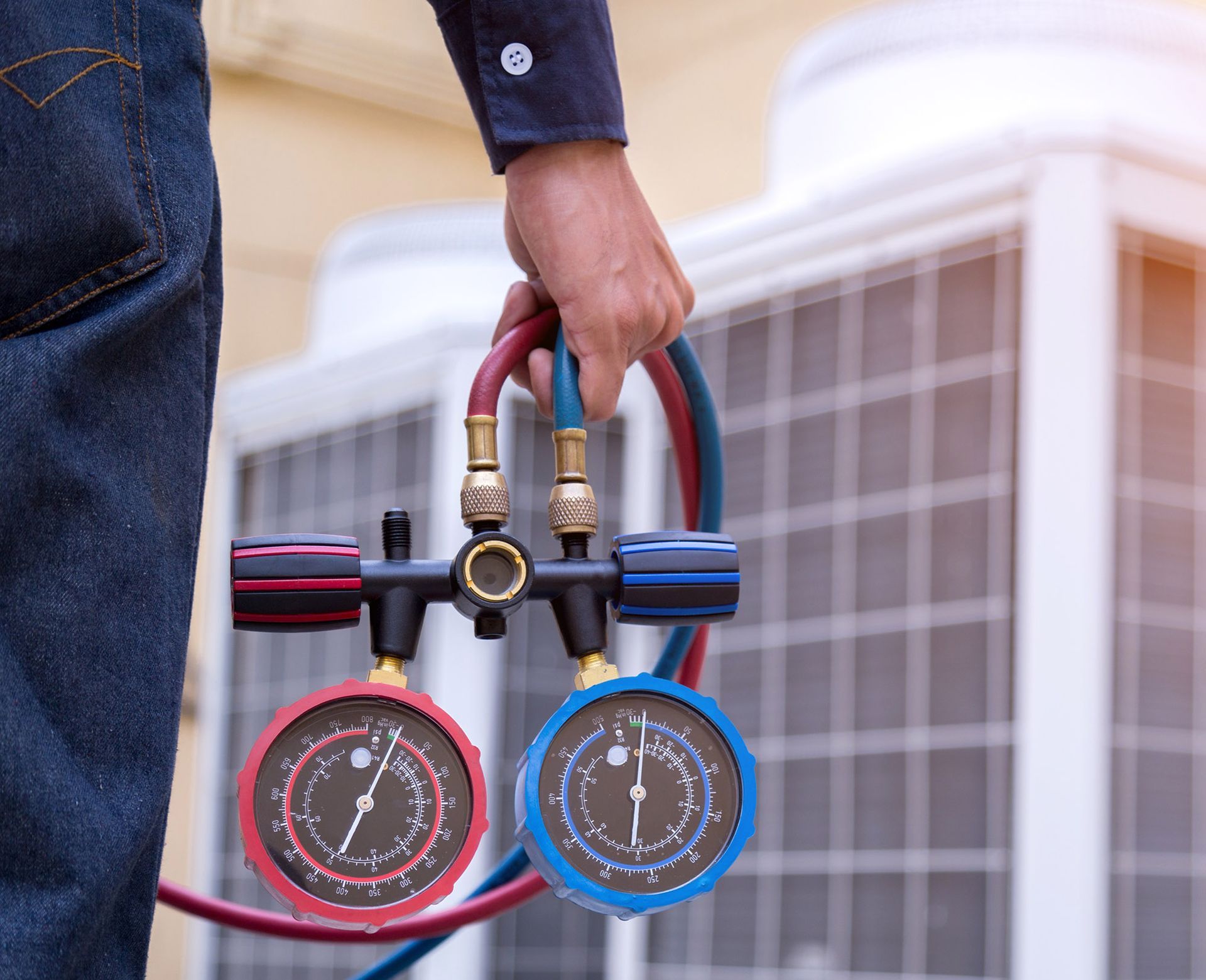 Person holding a gauge manifold, standing near an air conditioning unit.