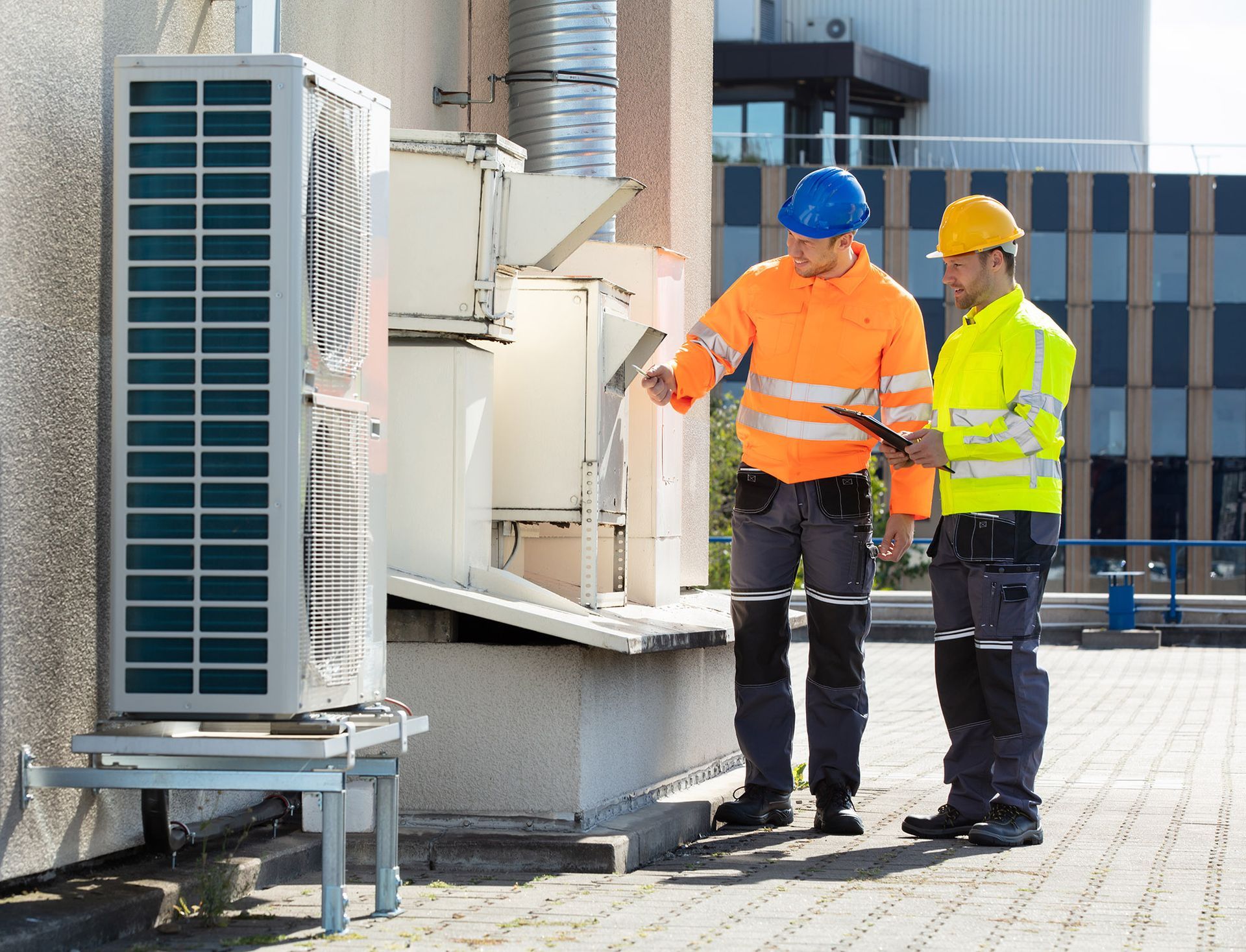 Two HVAC technicians examining equipment on a rooftop. One points, the other holds a clipboard.