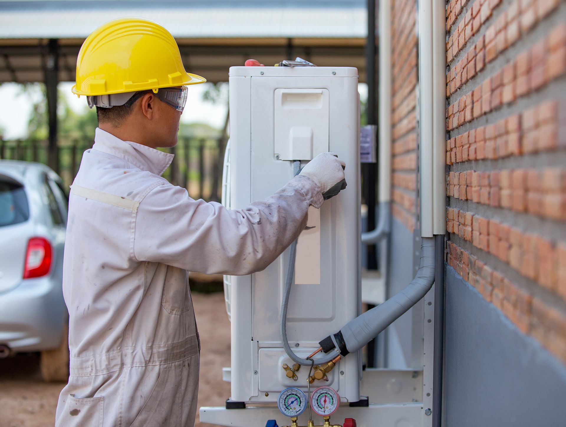 HVAC technician in yellow hard hat, working on an air conditioning unit attached to a brick wall.