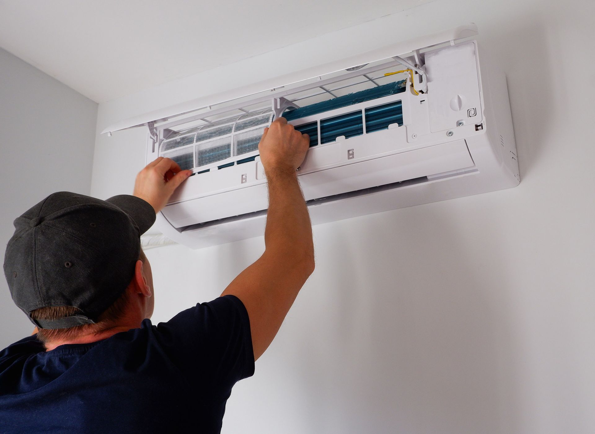 Person in cap cleaning air conditioner filter on white wall.