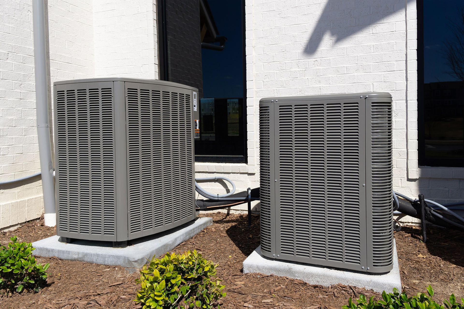 Two gray air conditioning units on concrete pads beside a building.