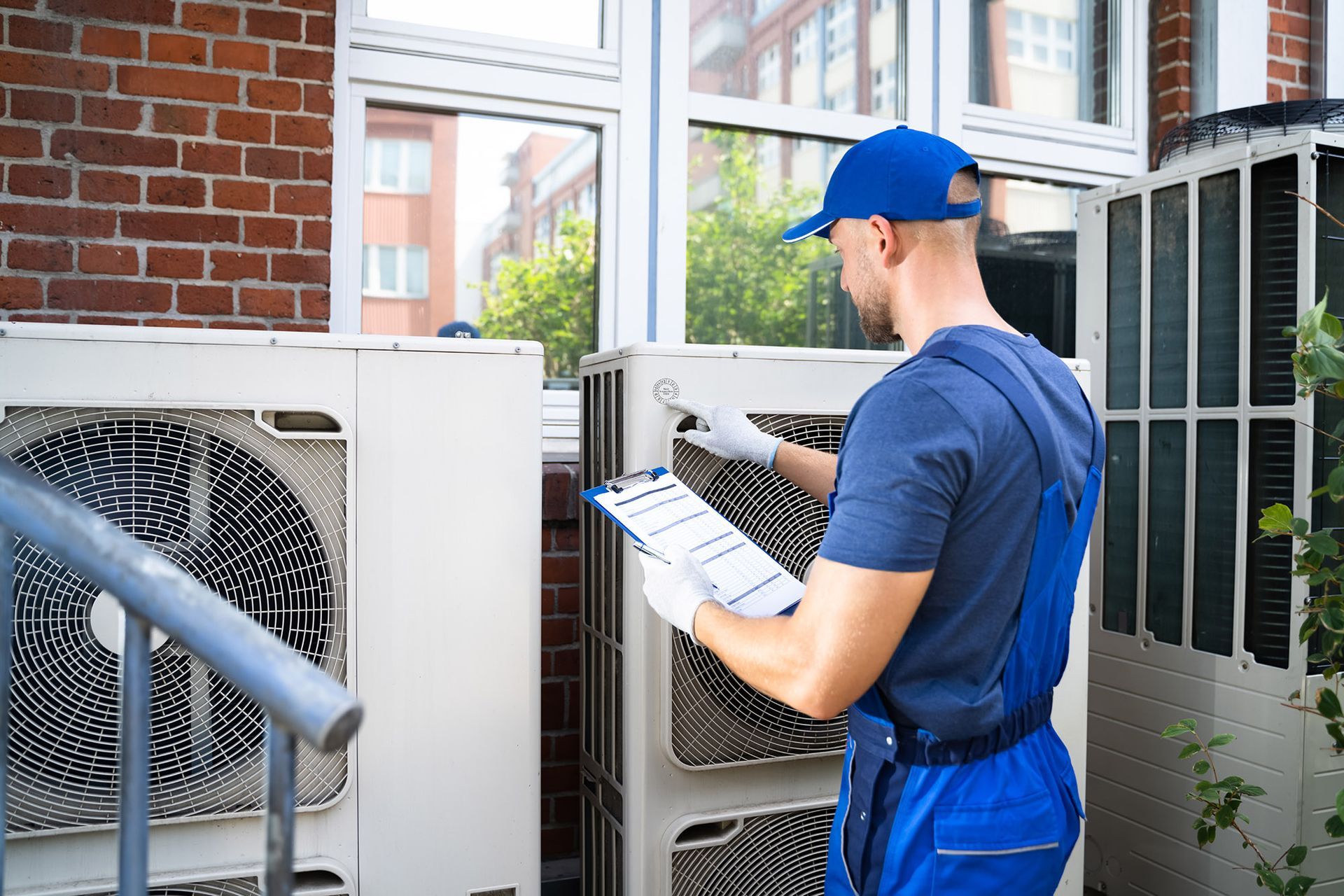 HVAC technician in blue overalls inspecting outdoor air conditioning units near a building, holding a clipboard.