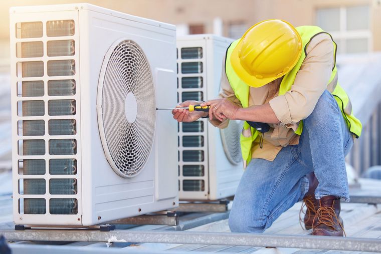 HVAC technician kneels, wearing hard hat and safety vest, repairing an outdoor air conditioning unit.