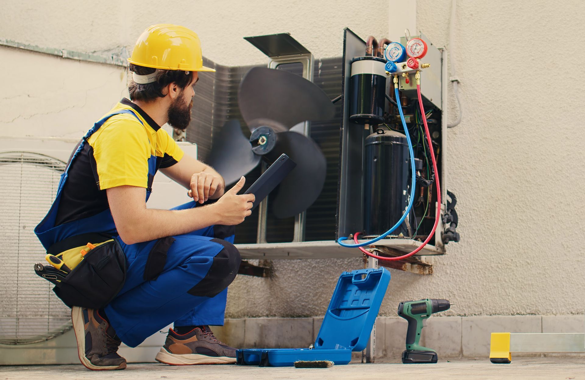 HVAC technician in yellow helmet and blue jumpsuit inspecting an outdoor air conditioning unit.