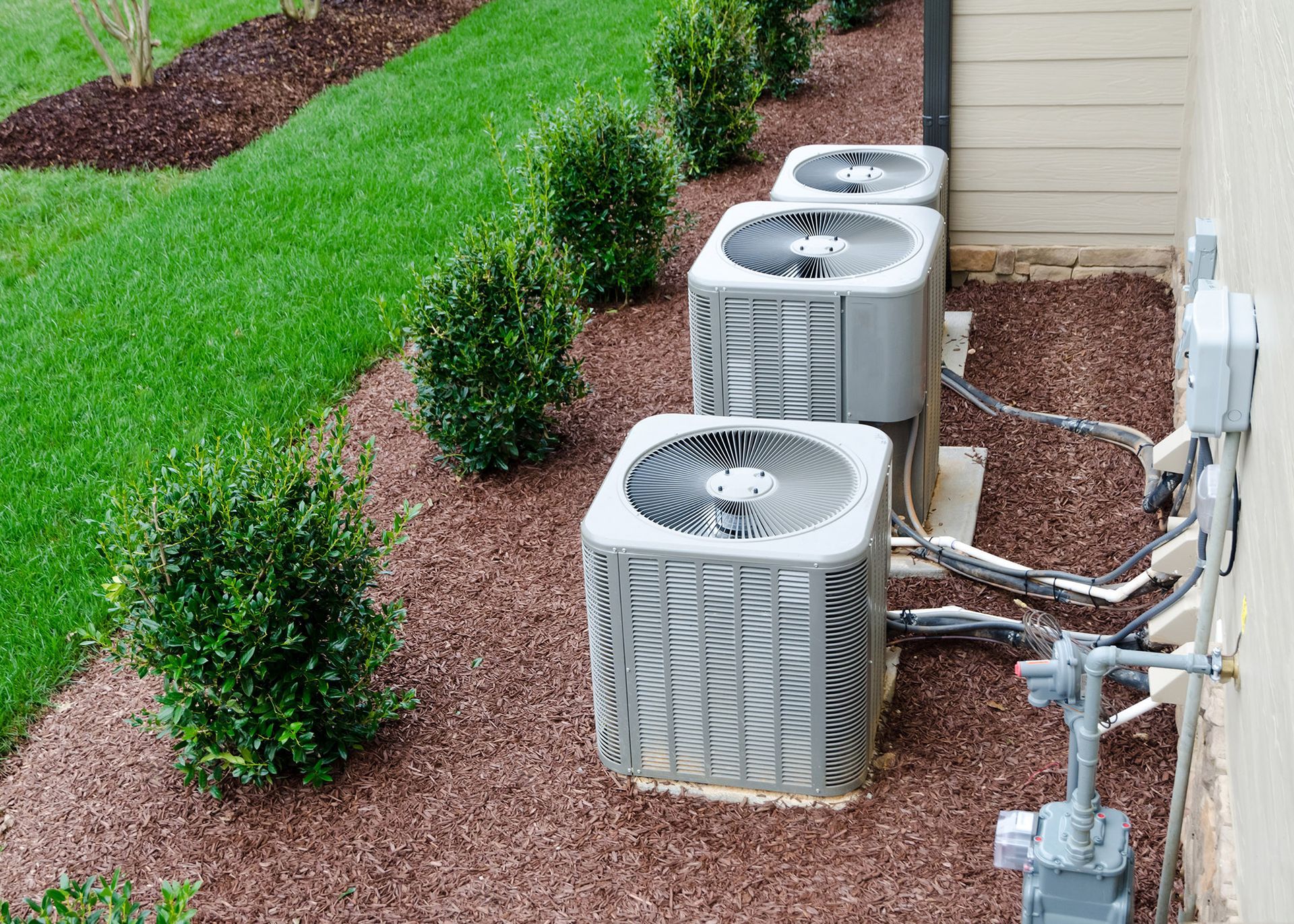 Air conditioning units lined up on a bed of mulch next to a building.
