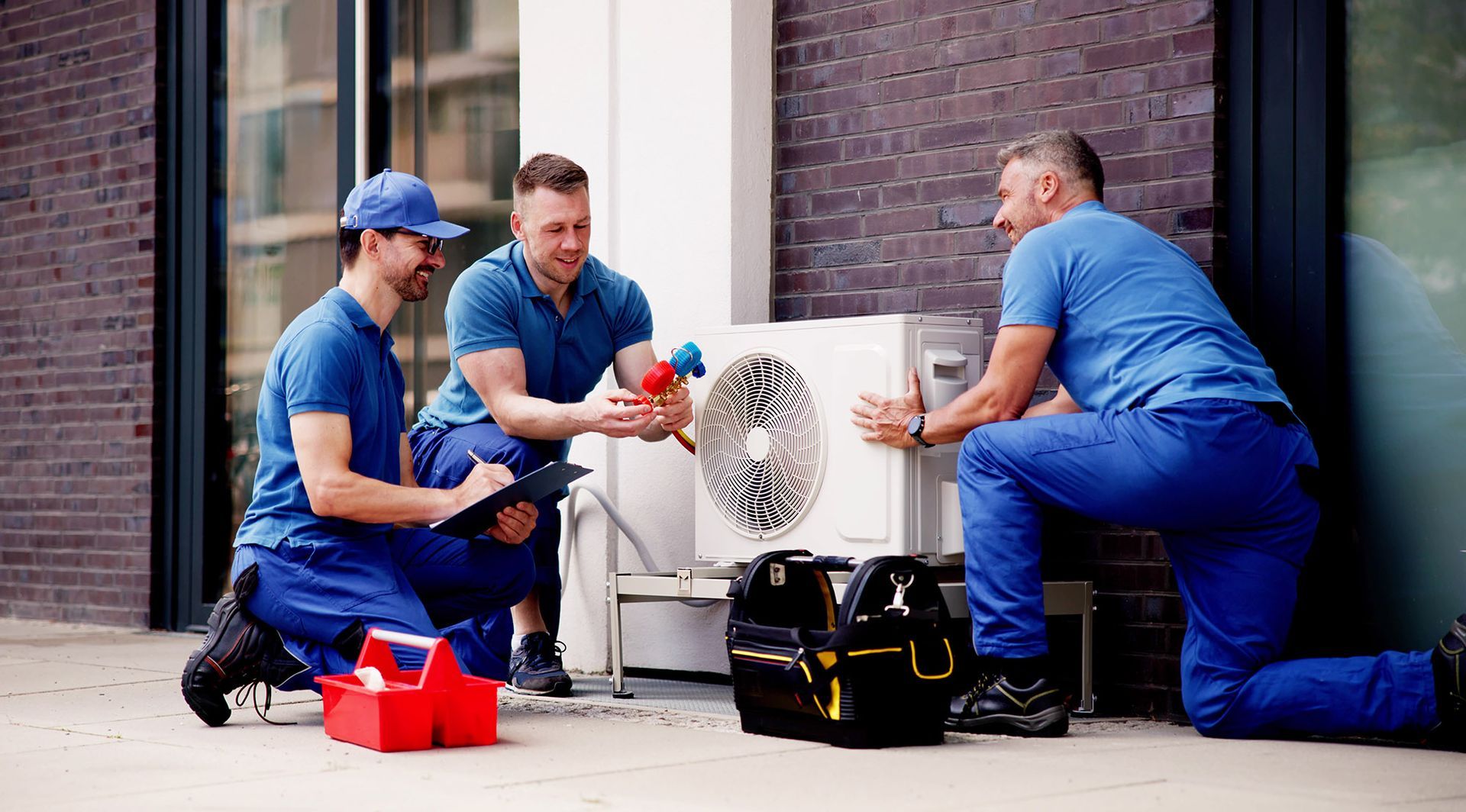 Three HVAC technicians in blue uniforms work on an outdoor unit; one kneels, two squat.