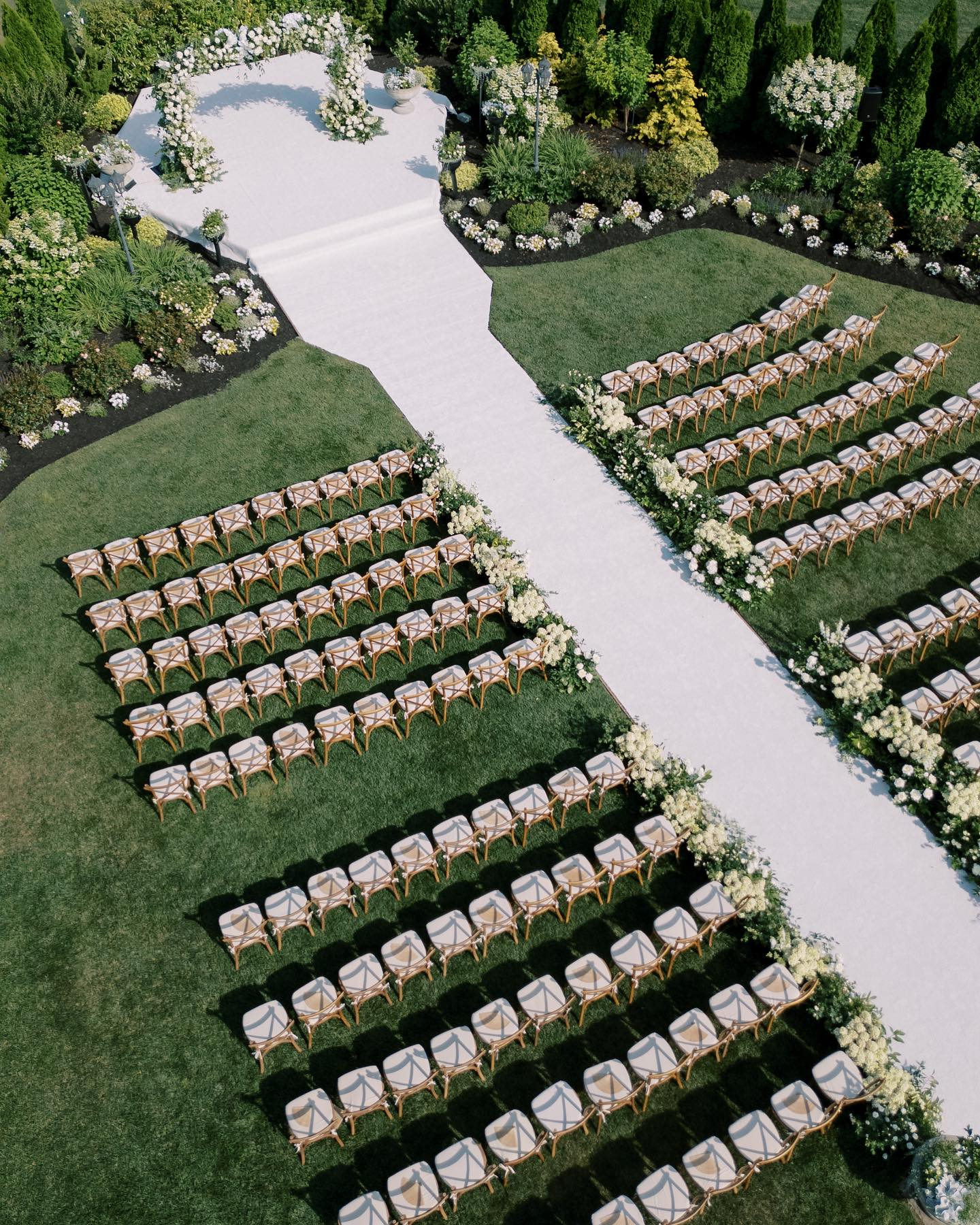 Rows of white chairs arranged in a garden ceremony setup beside a central aisle and floral arch