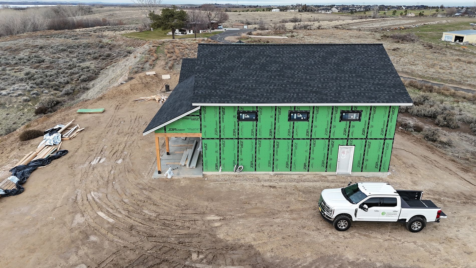 House under construction with green wrap, black roof, and white truck parked nearby on dirt lot.