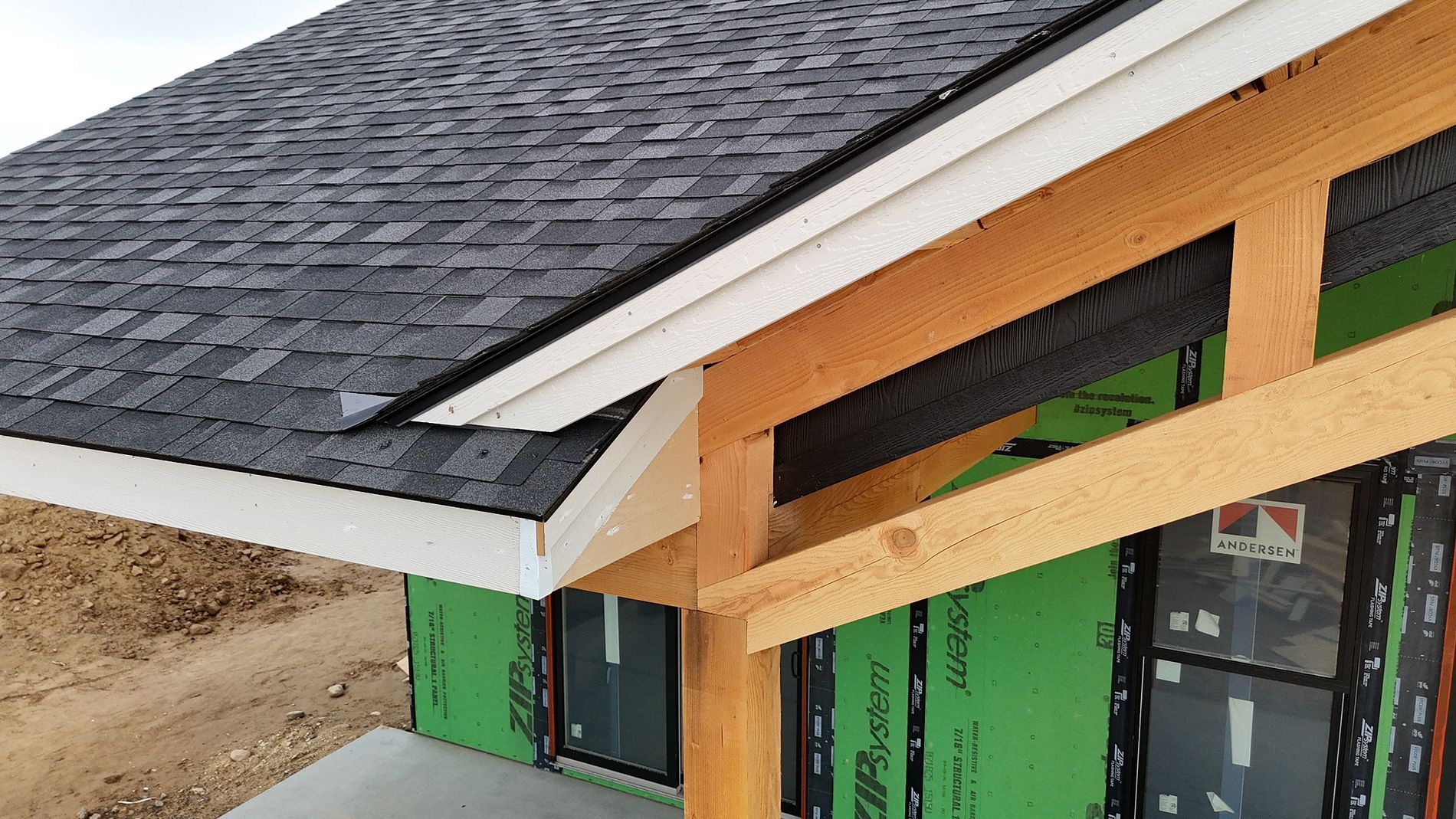 Construction of a house with black shingle roof, wood beams, white trim, and green wrap.