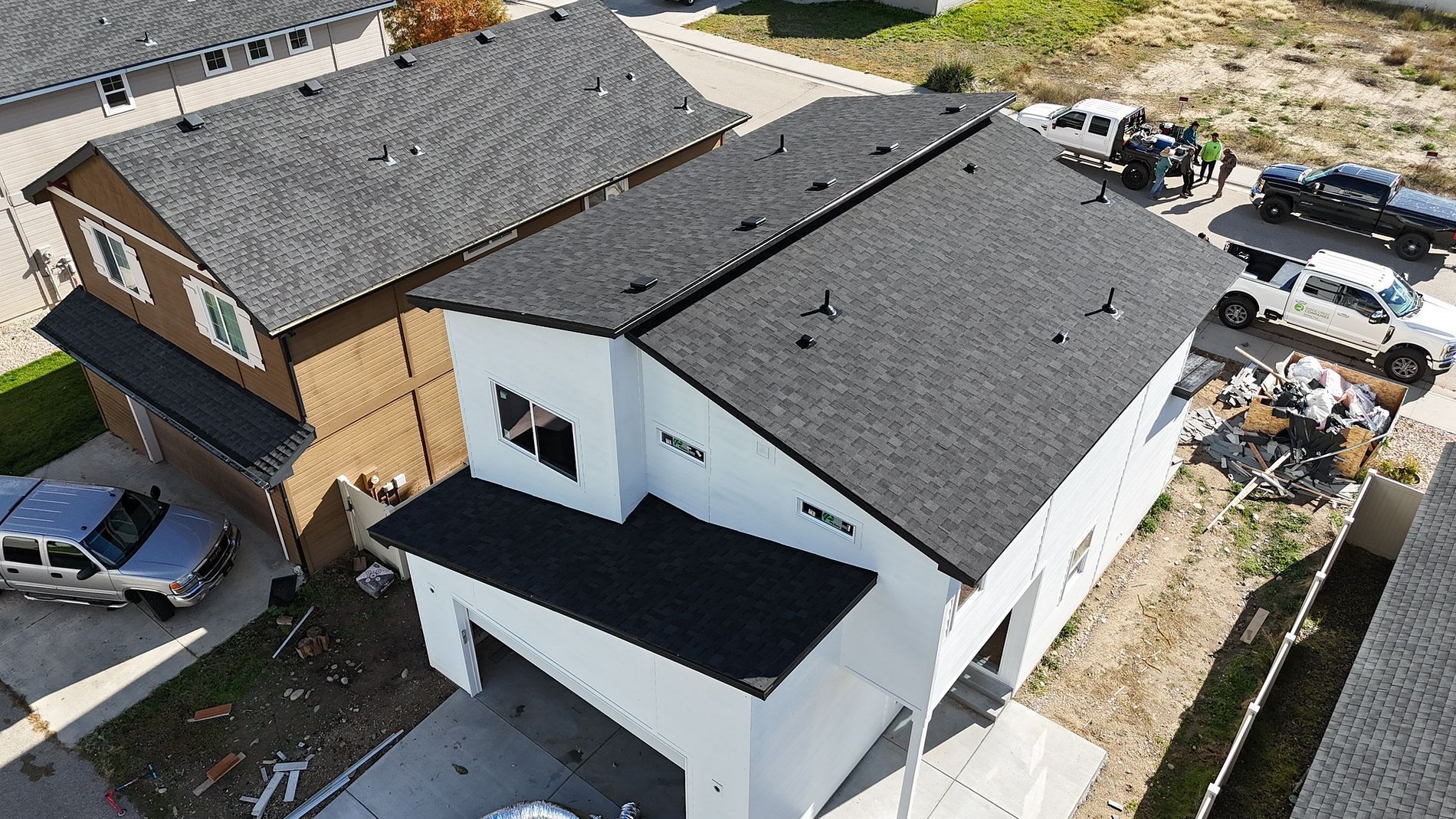 Aerial view of two-story houses, one with a new dark roof and white exterior, construction vehicles nearby.