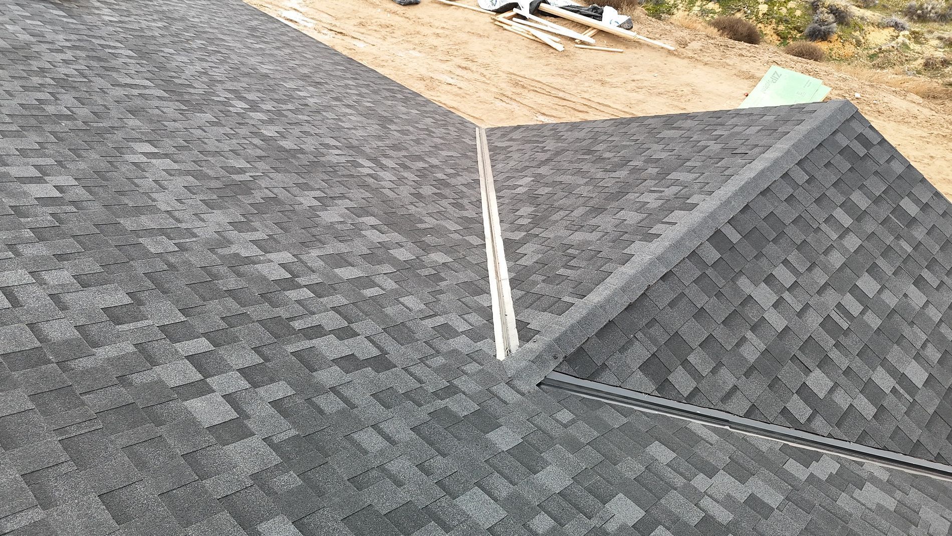 Overhead view of a roof with gray shingles. A portion of a structure and dirt are visible in the background.