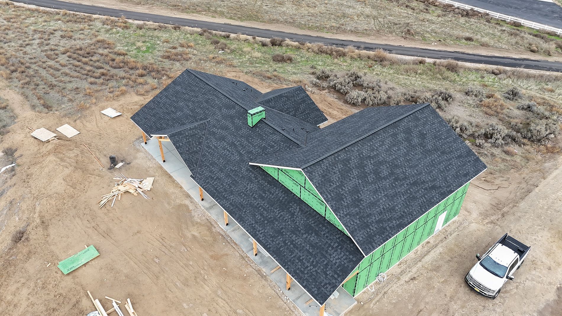 Aerial view of a house under construction with a dark gray shingled roof, green wrap, and a white pickup truck parked nearby.