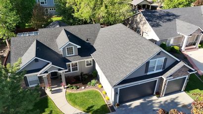 Aerial view of a gray house with a dark roof and a two-car garage, surrounded by green grass and trees.