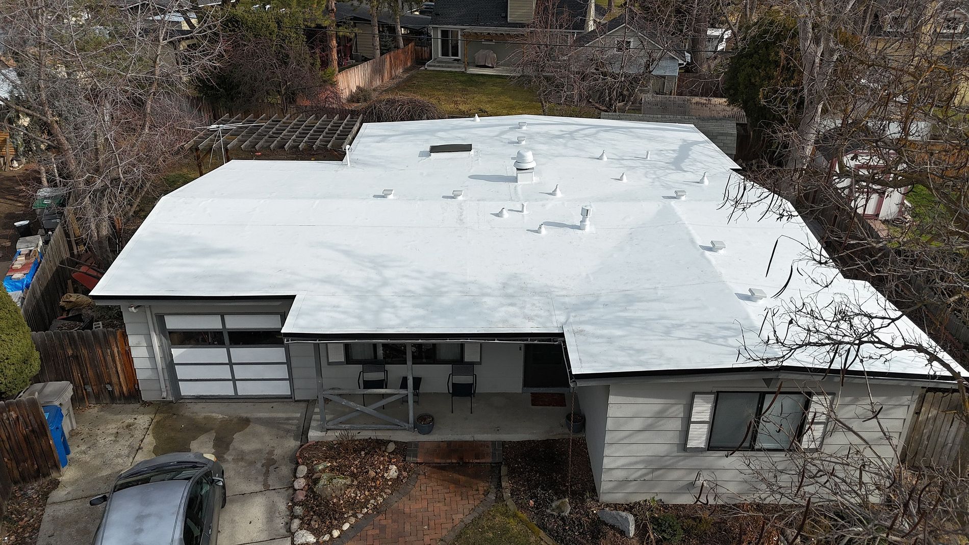 Overhead view of a one-story house with a white roof and garage door; bare trees surround.