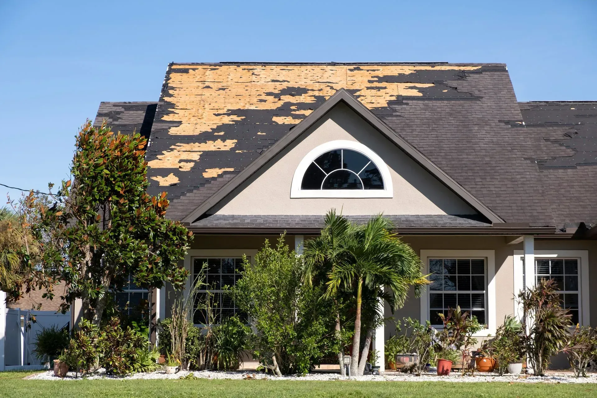 House with damaged roof, blue sky, and green lawn.  Patchy shingles visible.