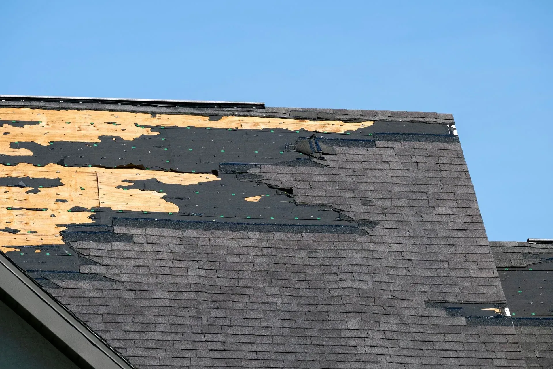 Damaged asphalt shingle roof with missing shingles and exposed underlayment against a blue sky.
