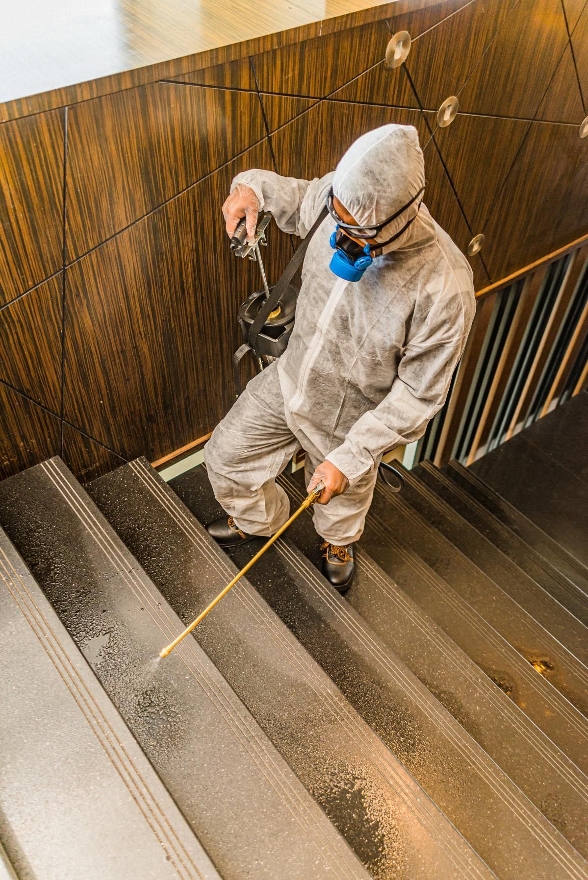 A worker in protective gear and a mask uses a sprayer to disinfect a set of indoor stairs.