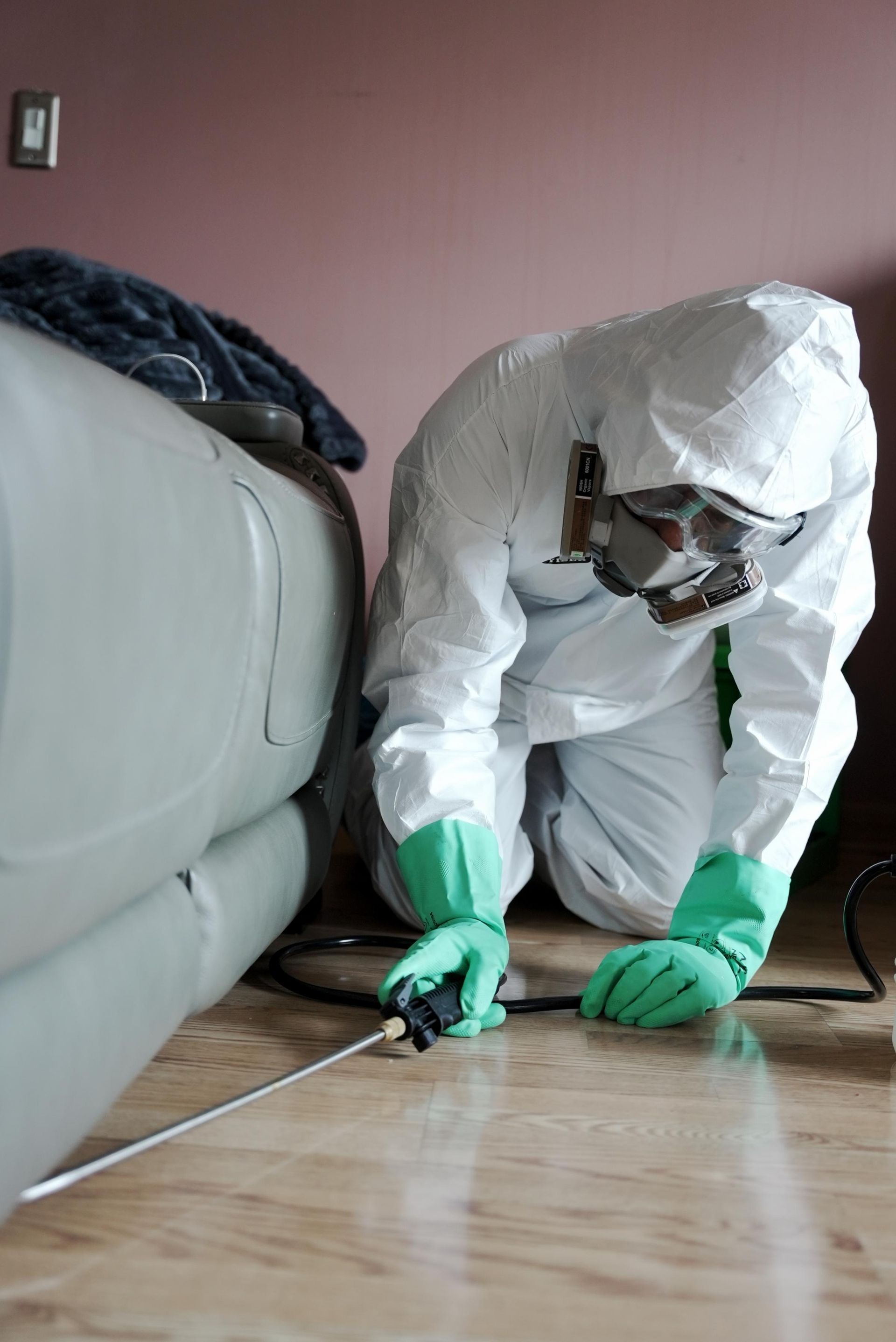 A pest control technician in a white hazmat suit and mask uses a sprayer to treat the floor near a couch.
