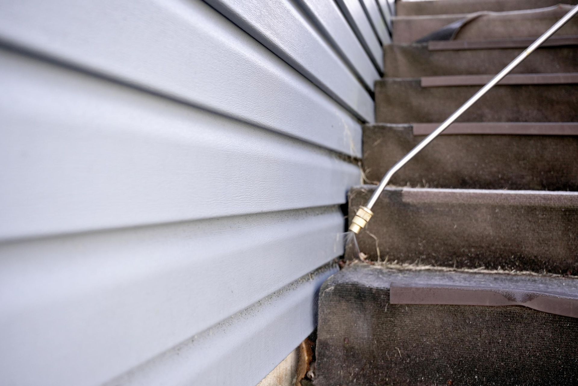 A pressure washer nozzle sprays water at the base of light gray vinyl siding next to a set of dark outdoor concrete stairs.