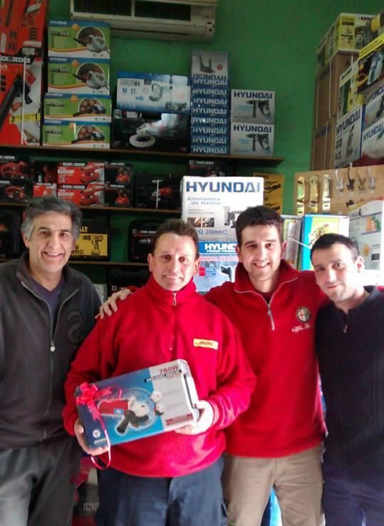 Cuatro hombres posan en una ferretería. Uno sostiene un regalo; herramientas en estantes detrás de ellos.