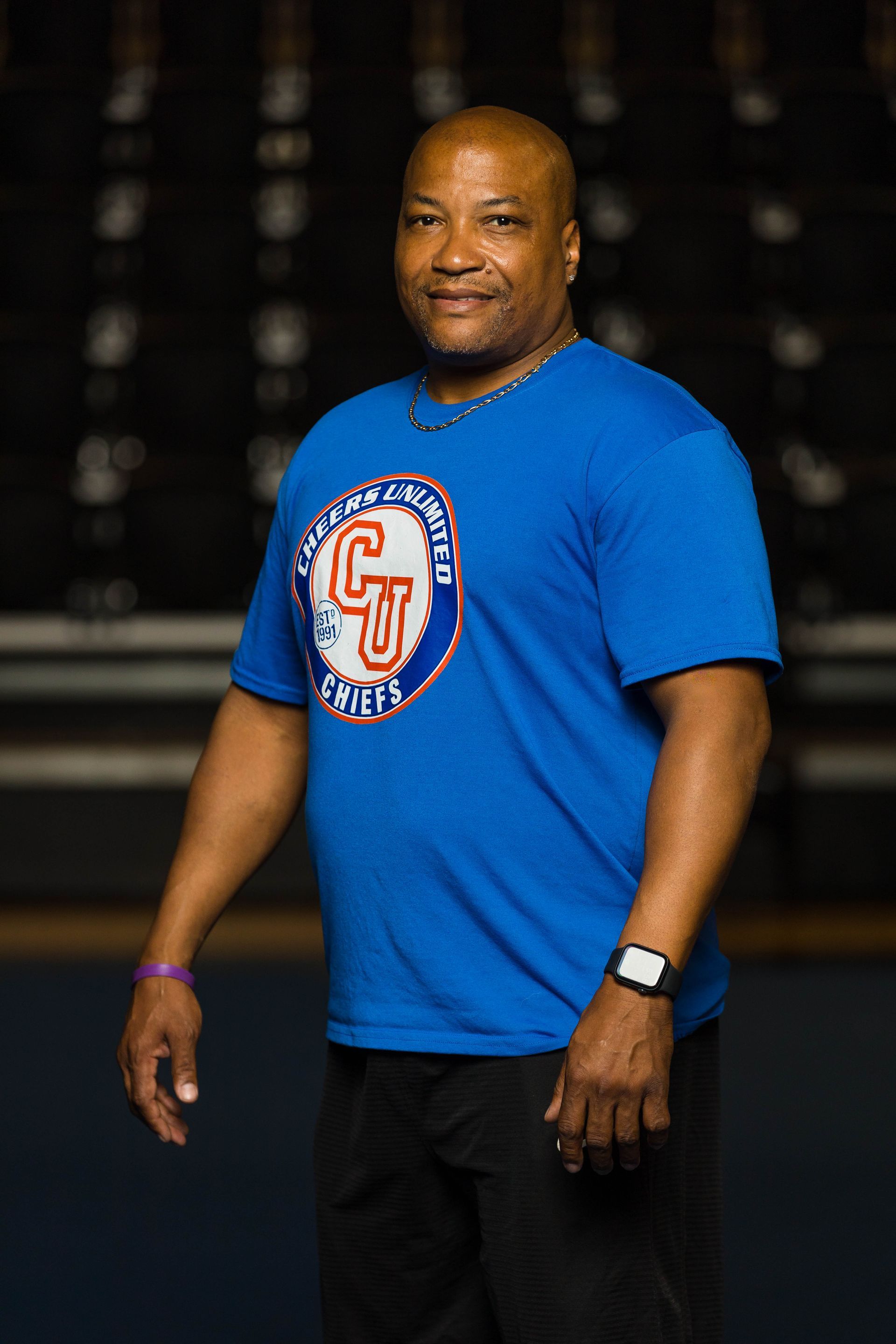 Man in blue t-shirt with logo, black pants, stands in an indoor setting.