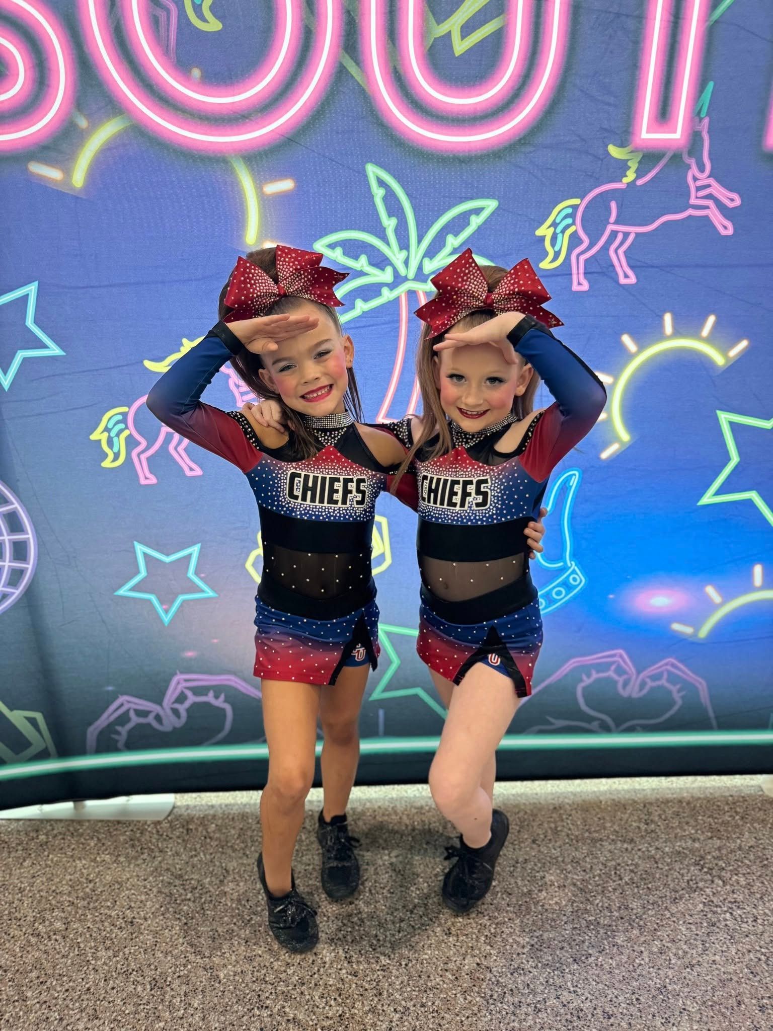Two young cheerleaders in red, white, and blue uniforms pose, smiling, hands on foreheads, in front of a colorful backdrop.