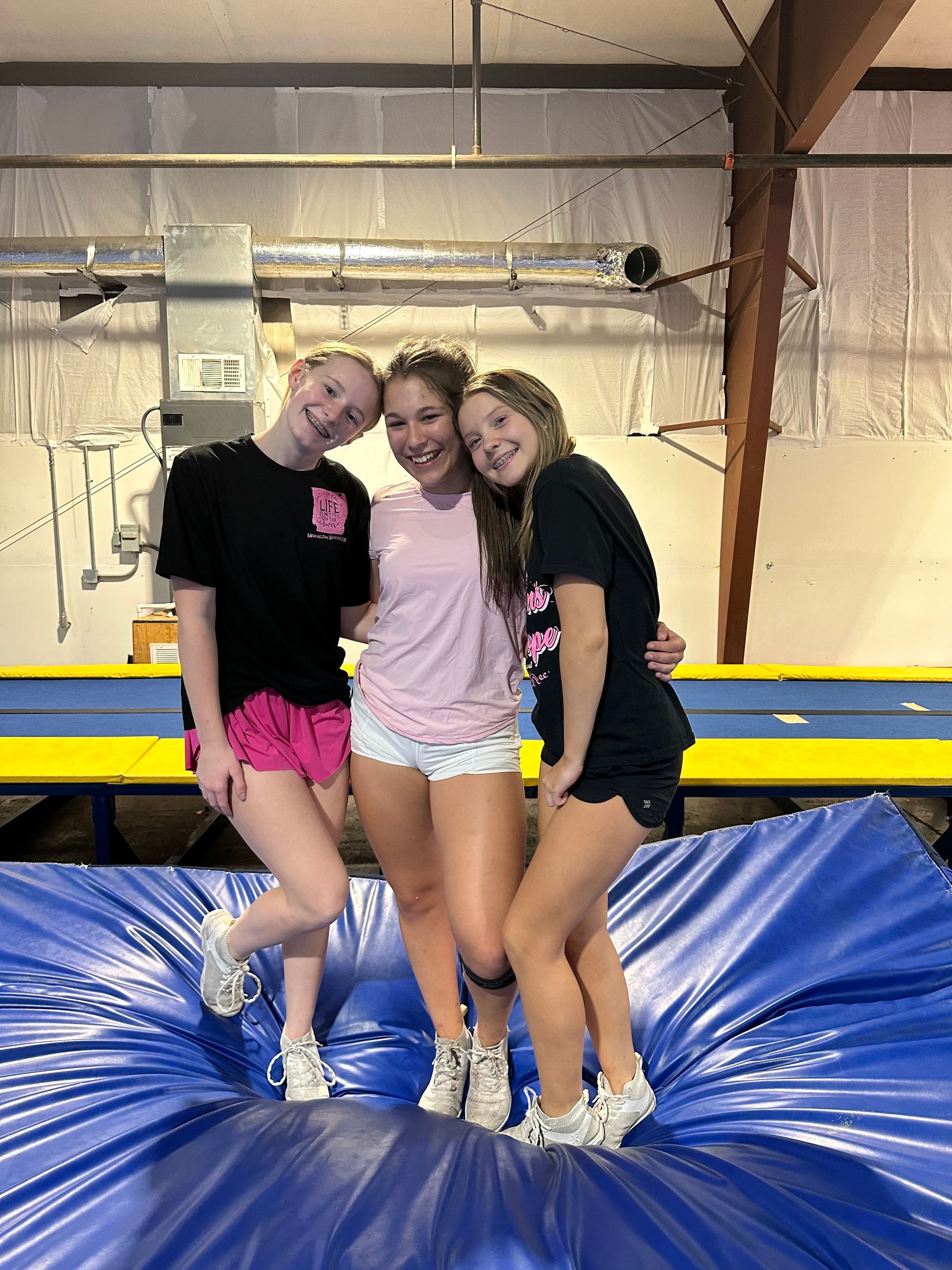 Three smiling girls standing on a blue mat, wearing athletic clothes in a gym.