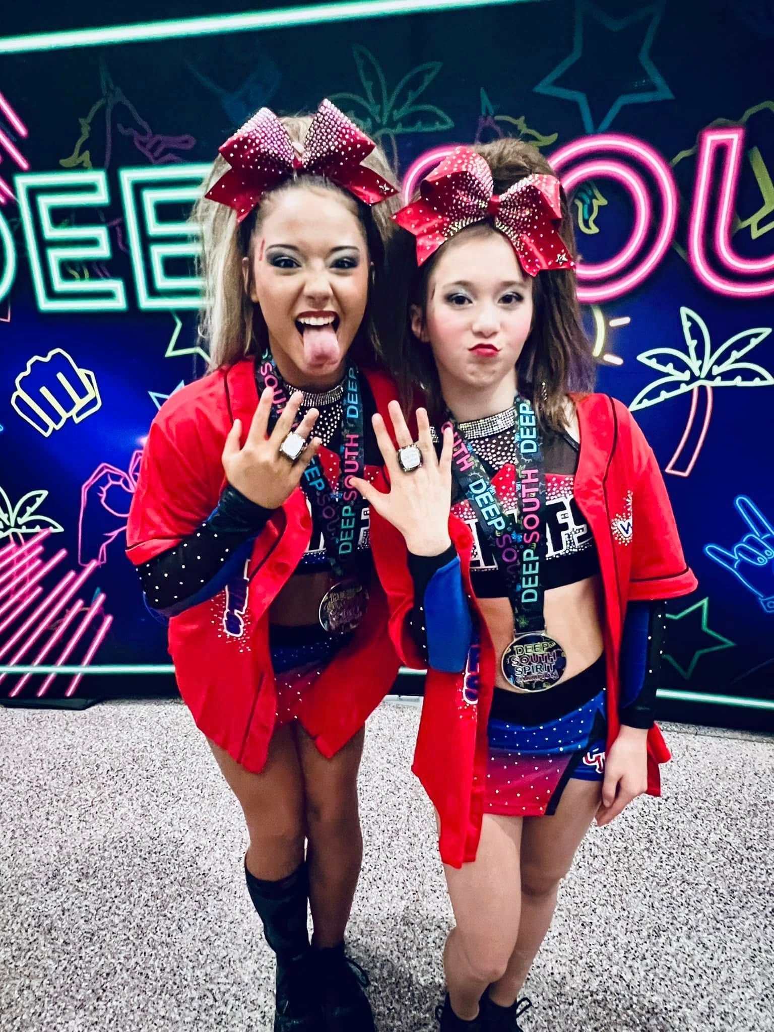 Two cheerleaders in red uniforms pose, showing off rings. They have big bows in their hair, medals, and are making faces.