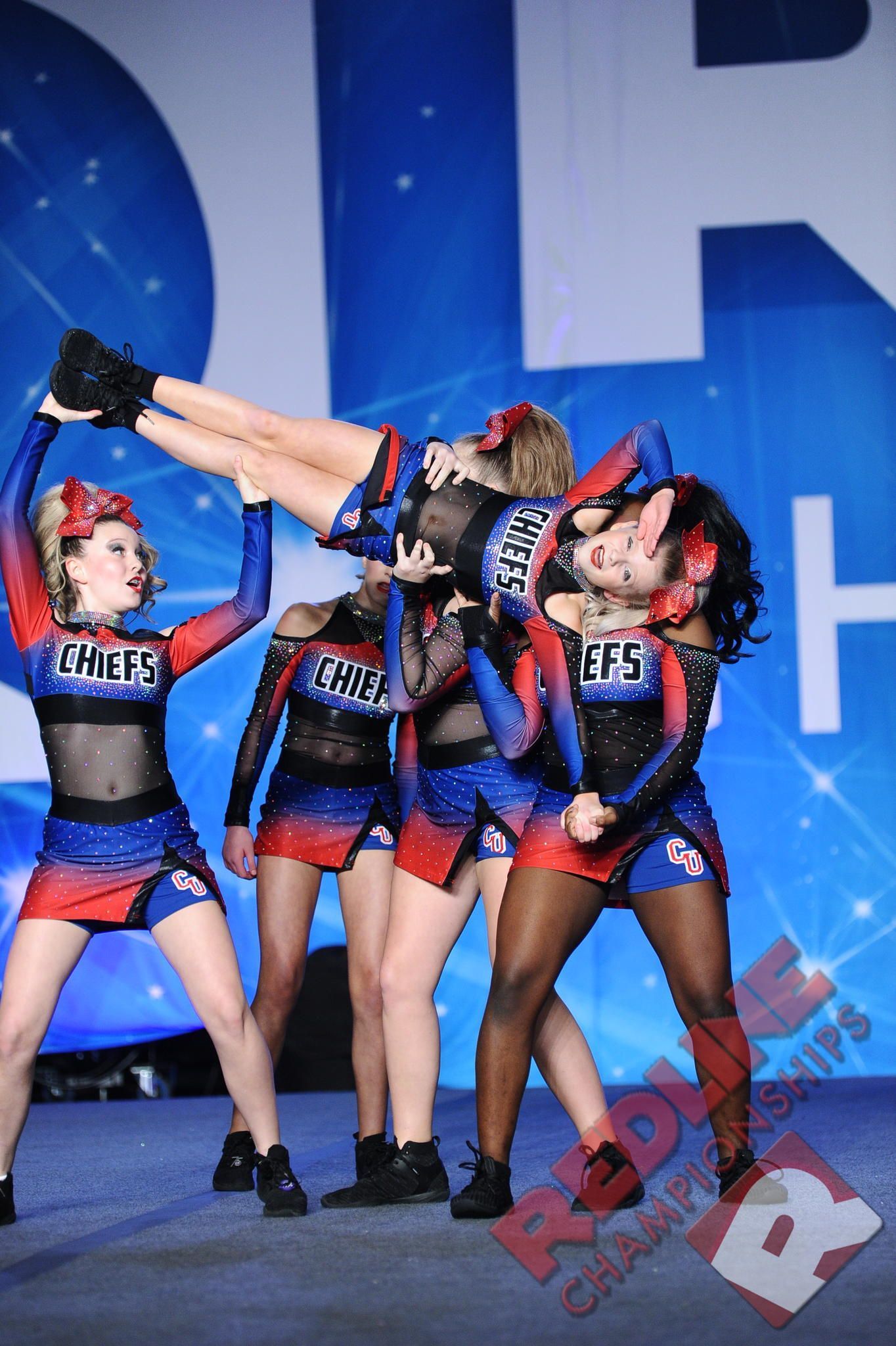 Cheerleaders in red and blue uniforms perform a pyramid stunt on a stage; backdrop features blue and white.