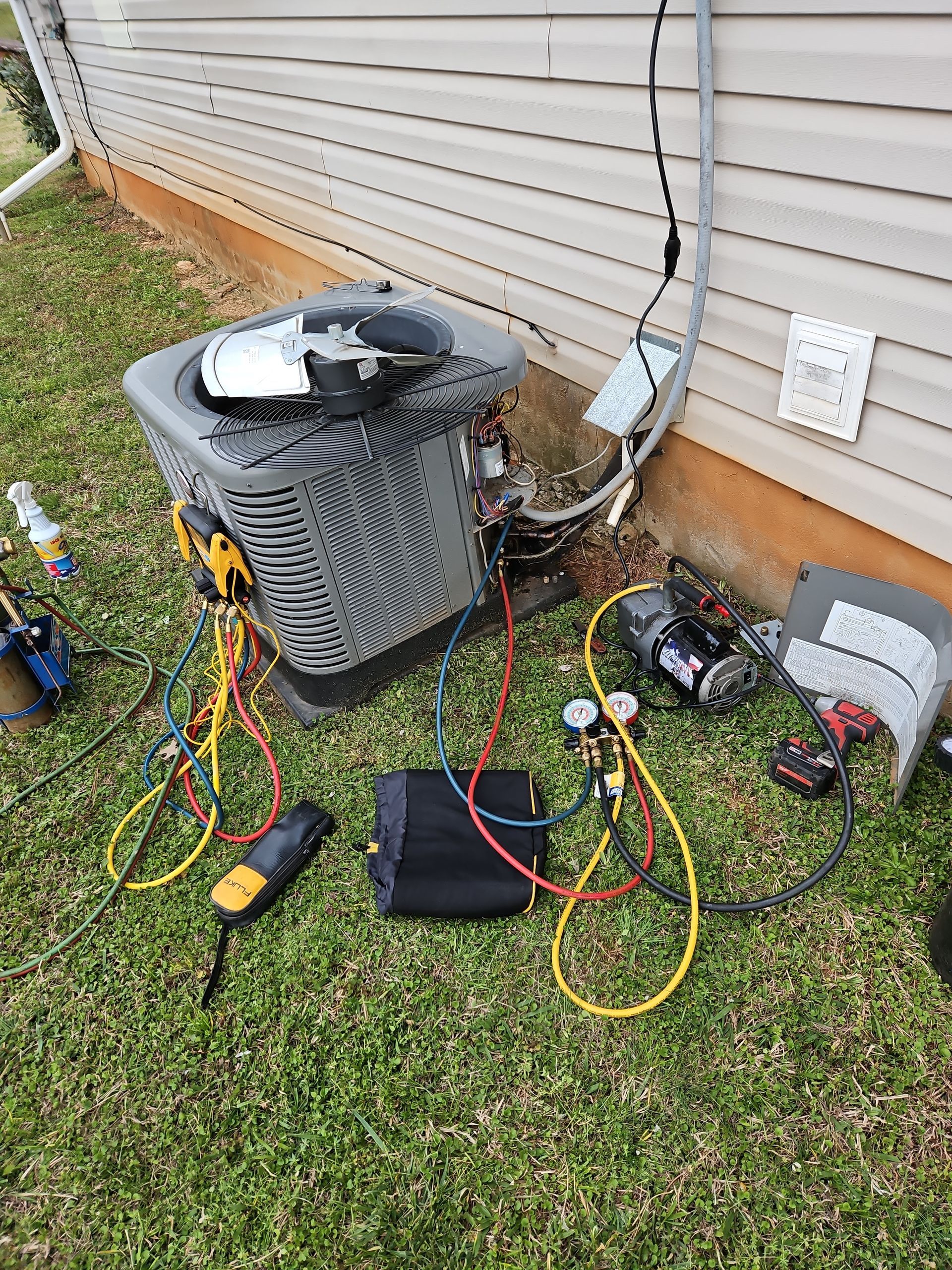 An air conditioning unit being serviced outdoors. Various tools and gauges are connected, with yellow, red, and blue hoses.