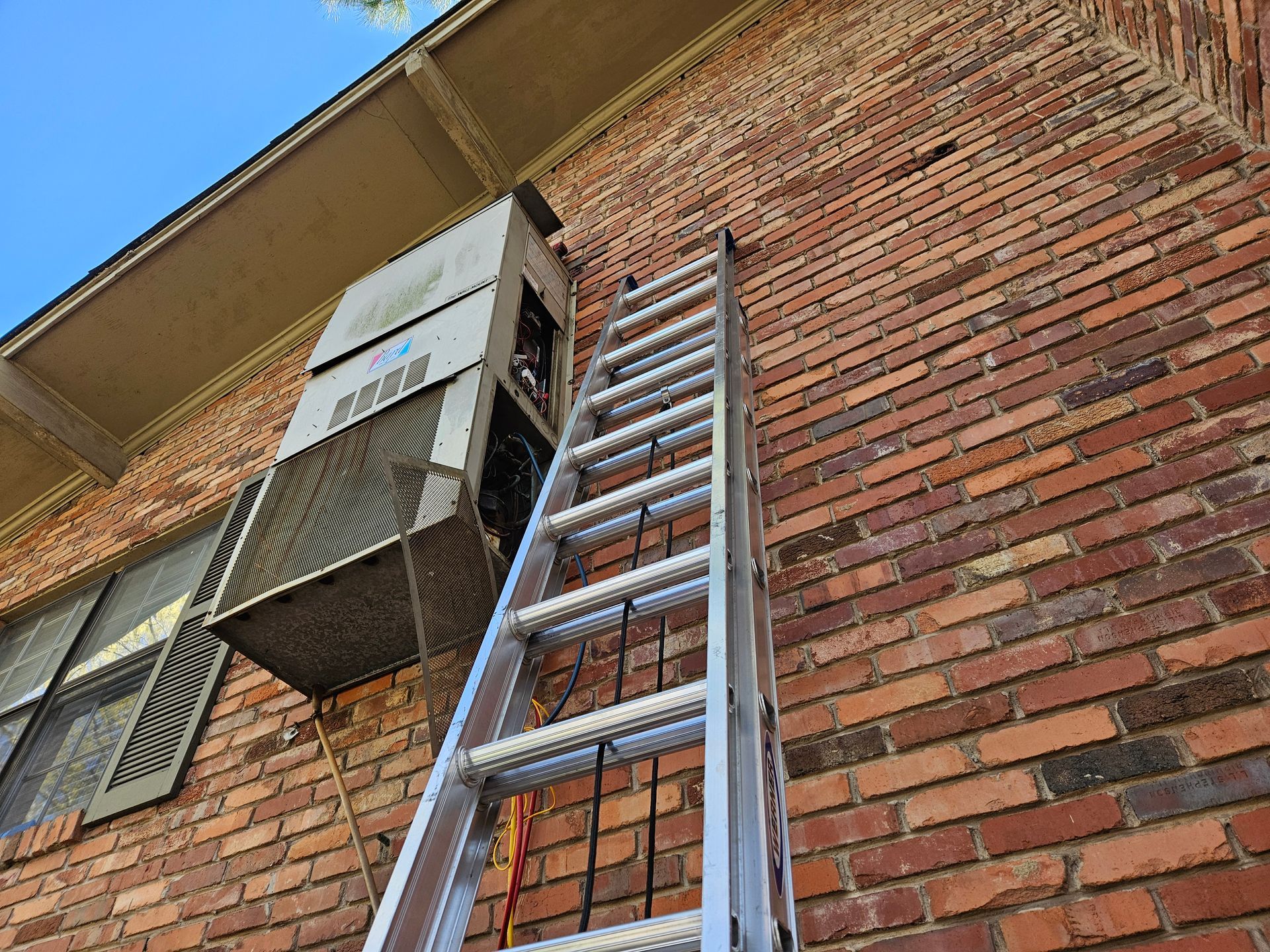 A metal ladder leans against a brick building next to a wall-mounted air conditioning unit.