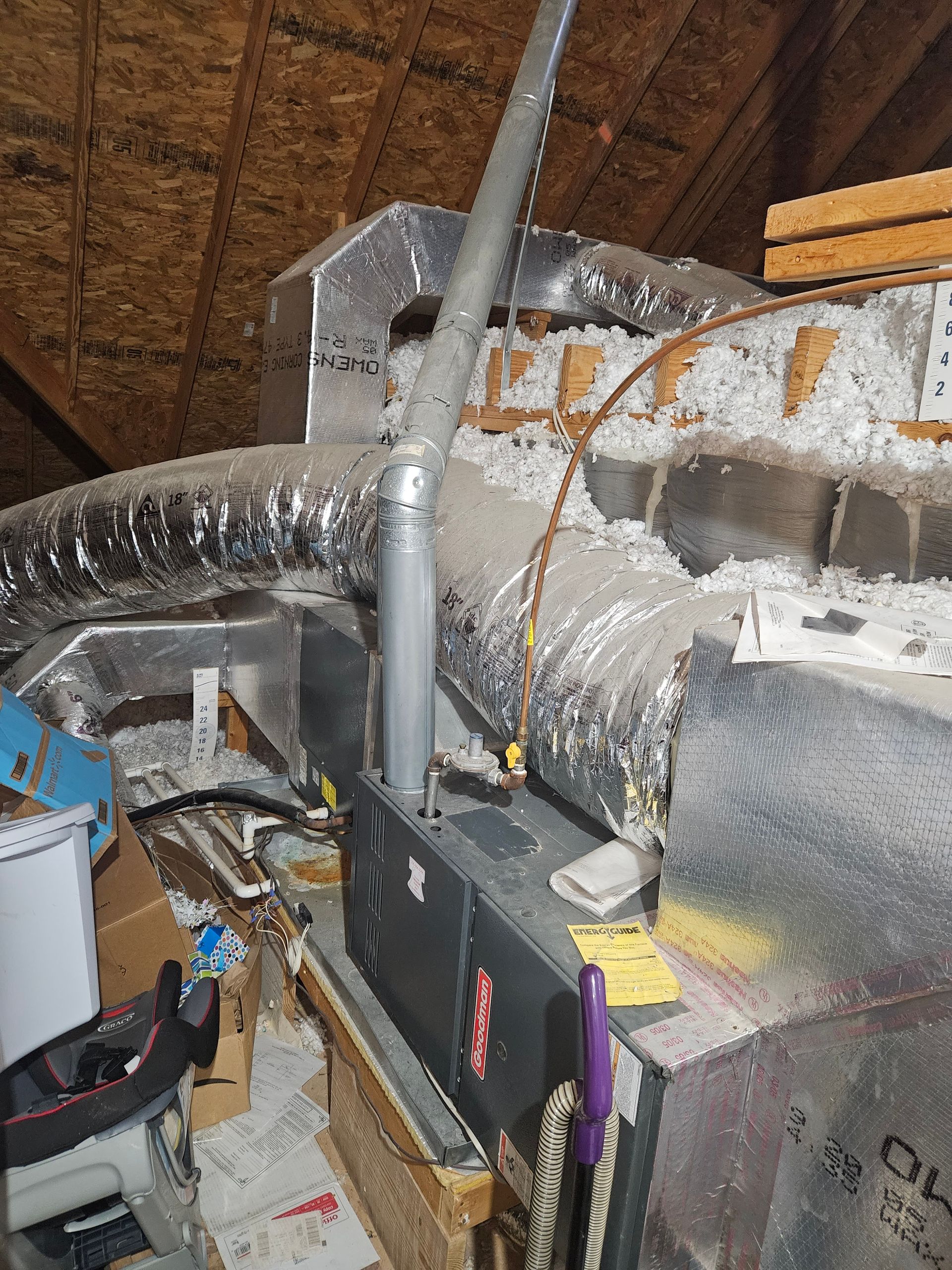HVAC unit in an attic. A silver flue pipe extends upwards from the dark-colored unit, surrounded by insulation and ductwork.