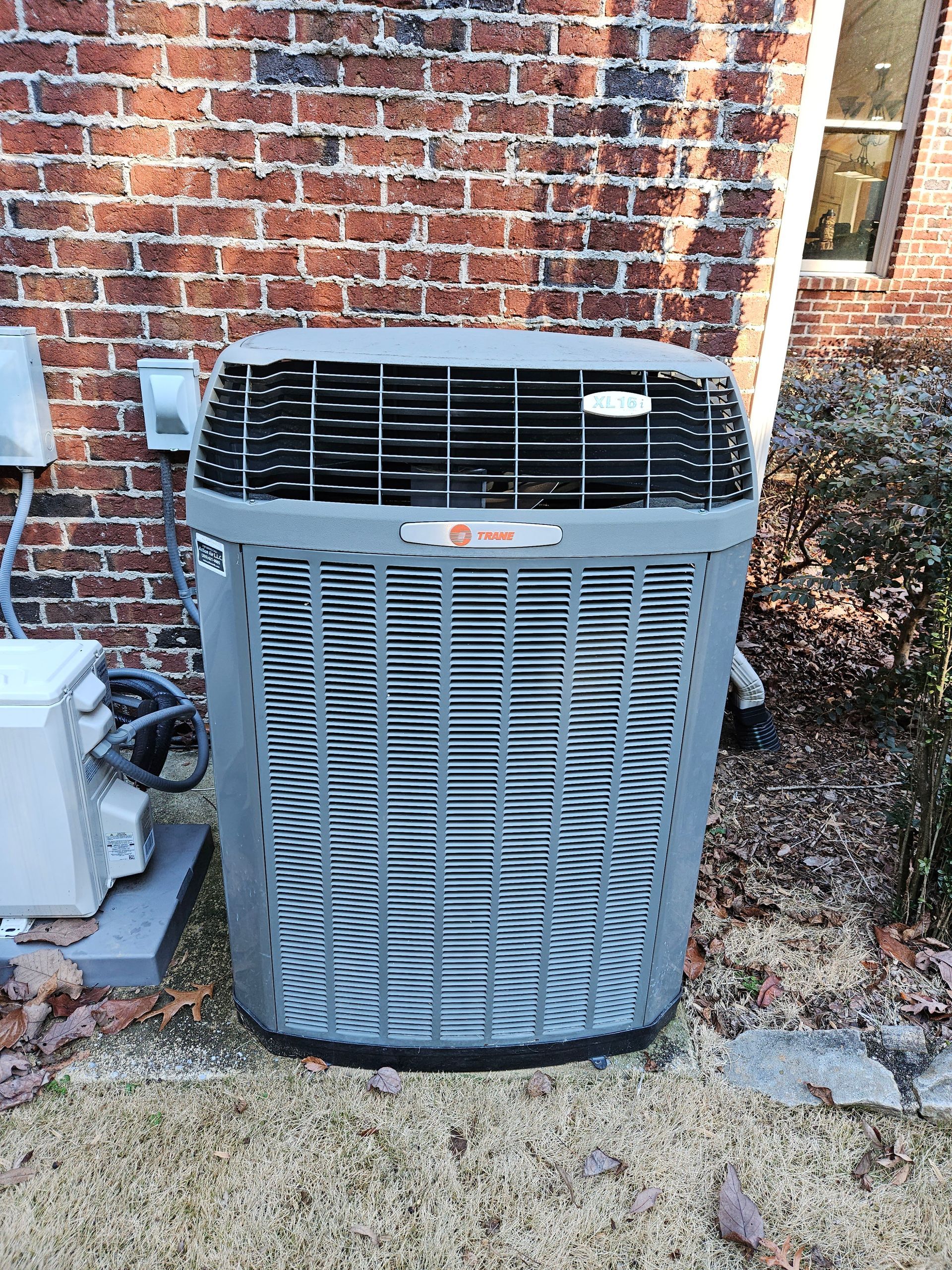 A Trane air conditioning unit against a brick wall. The unit is gray with a black top grill.