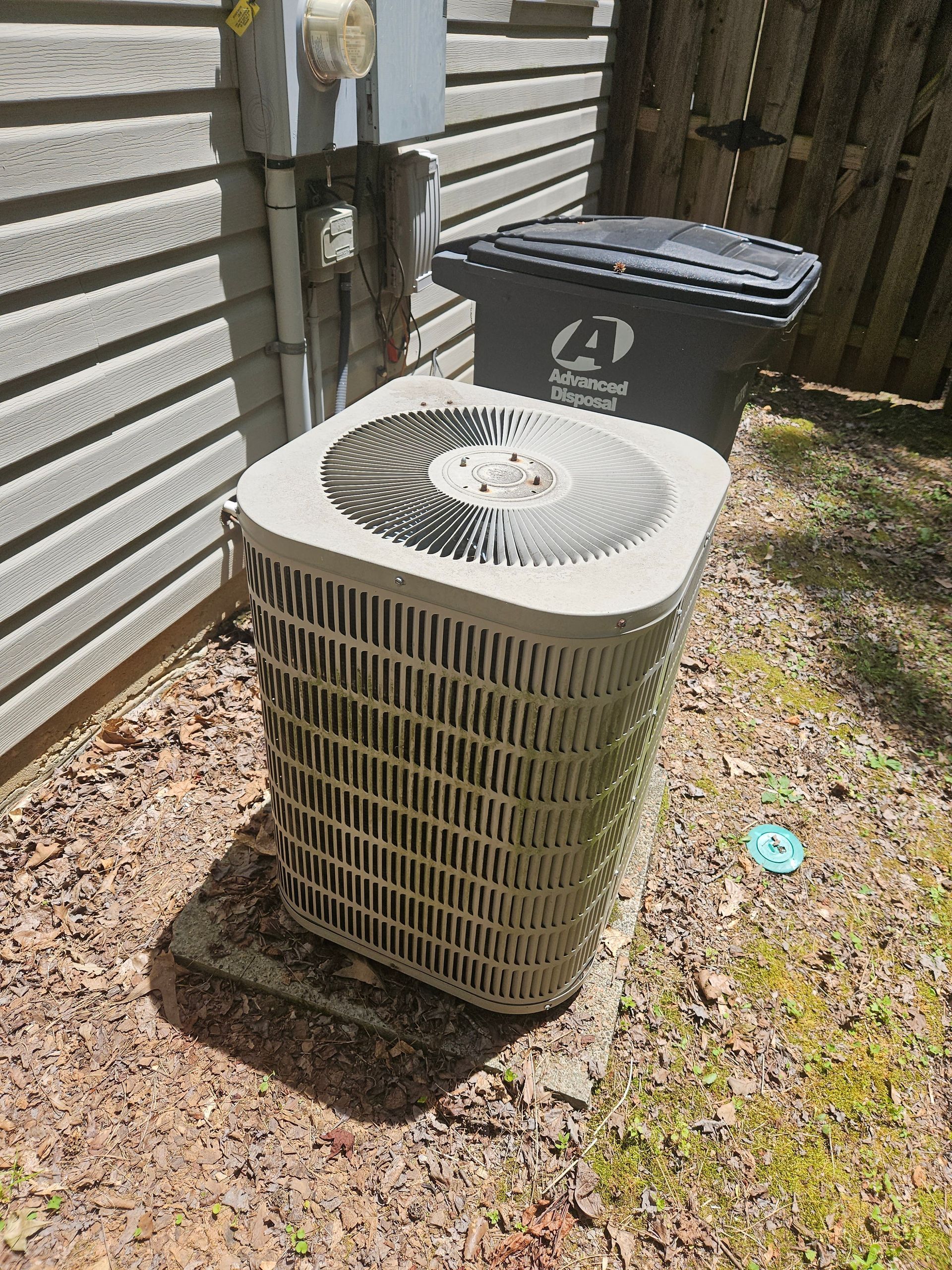 An outdoor air conditioning unit next to a trash bin and a building with siding. The unit is beige and gray.