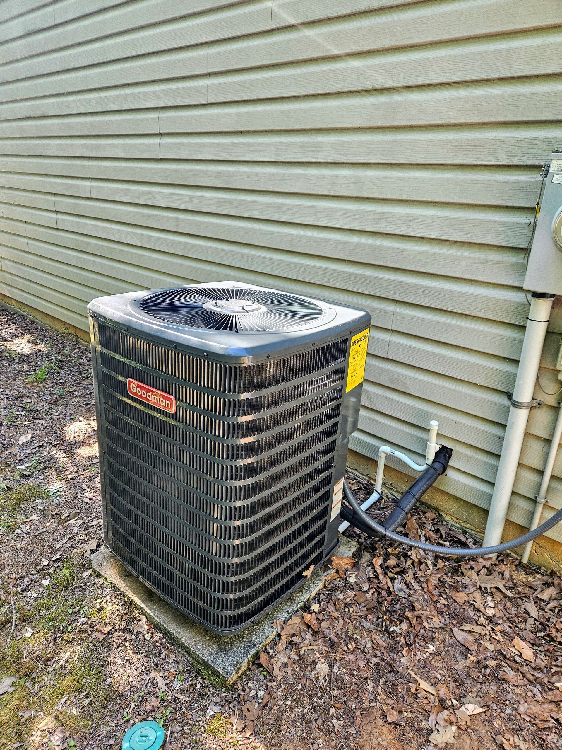 An outdoor air conditioning unit sits next to a light green sided house. The unit is black and mounted on a concrete pad.