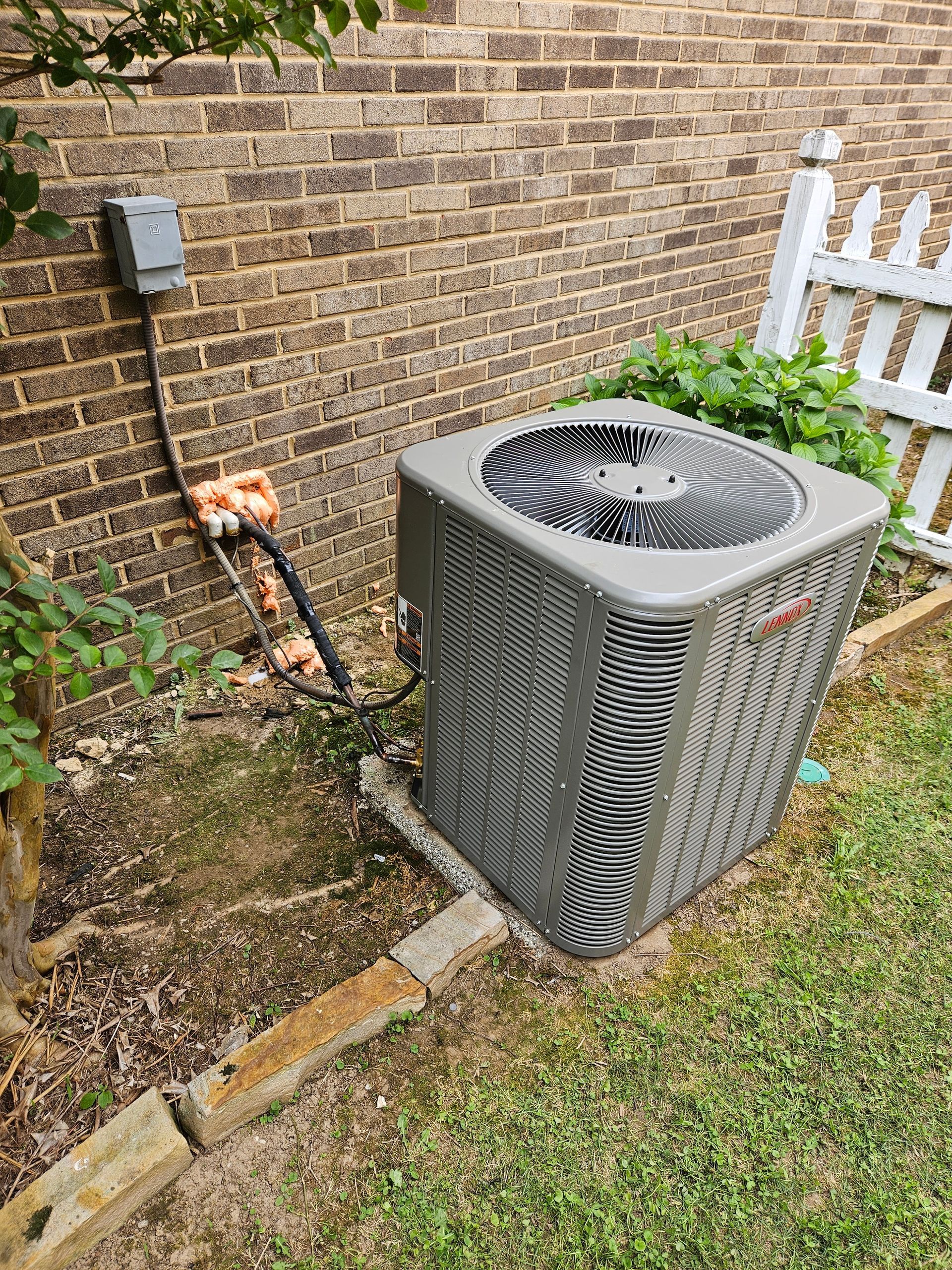An air conditioning unit sits near a brick wall and a white picket fence, with a small garden.