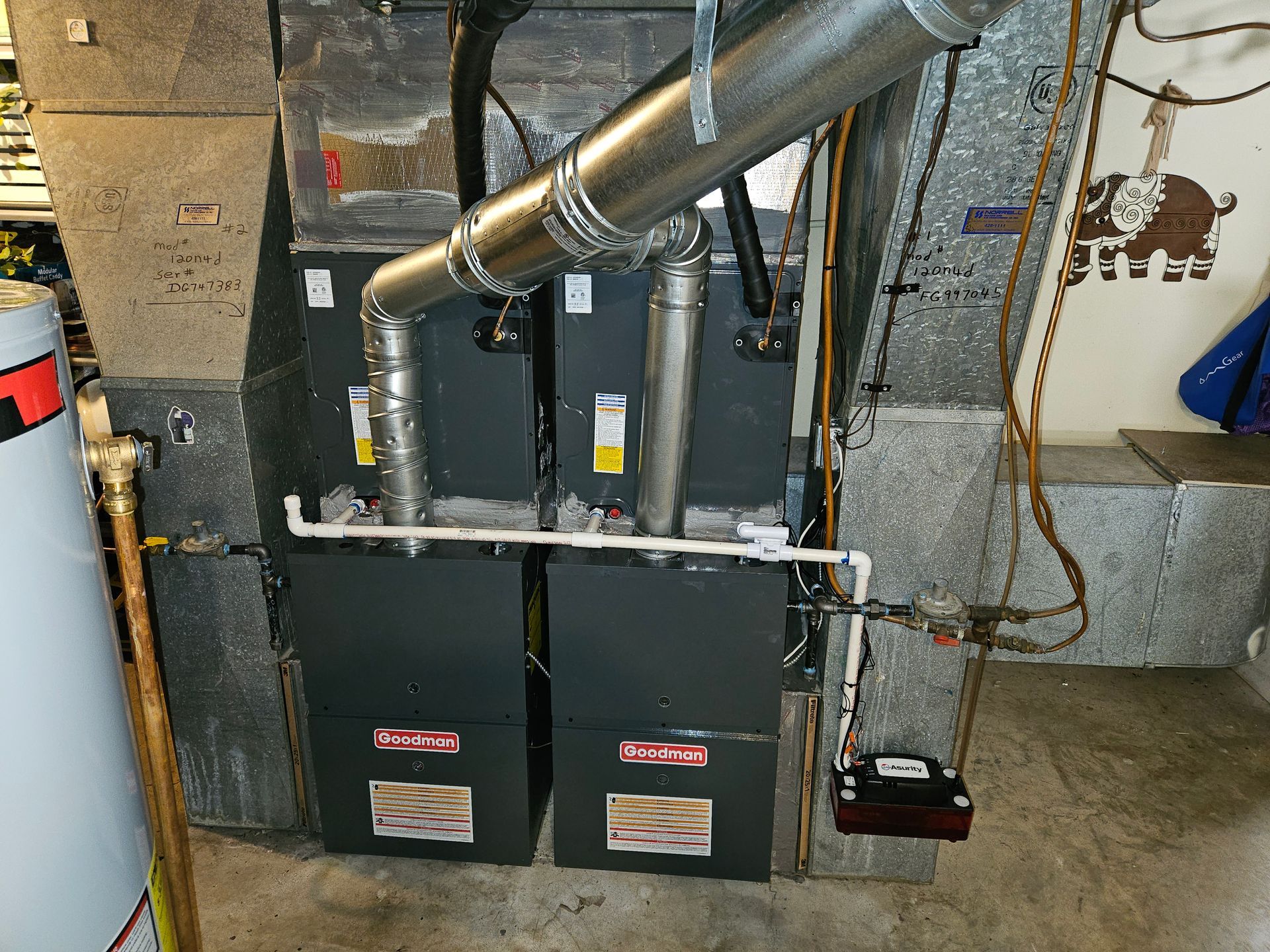 Two adjacent gray furnaces with silver ductwork in a basement setting, next to a water heater and wall decor.