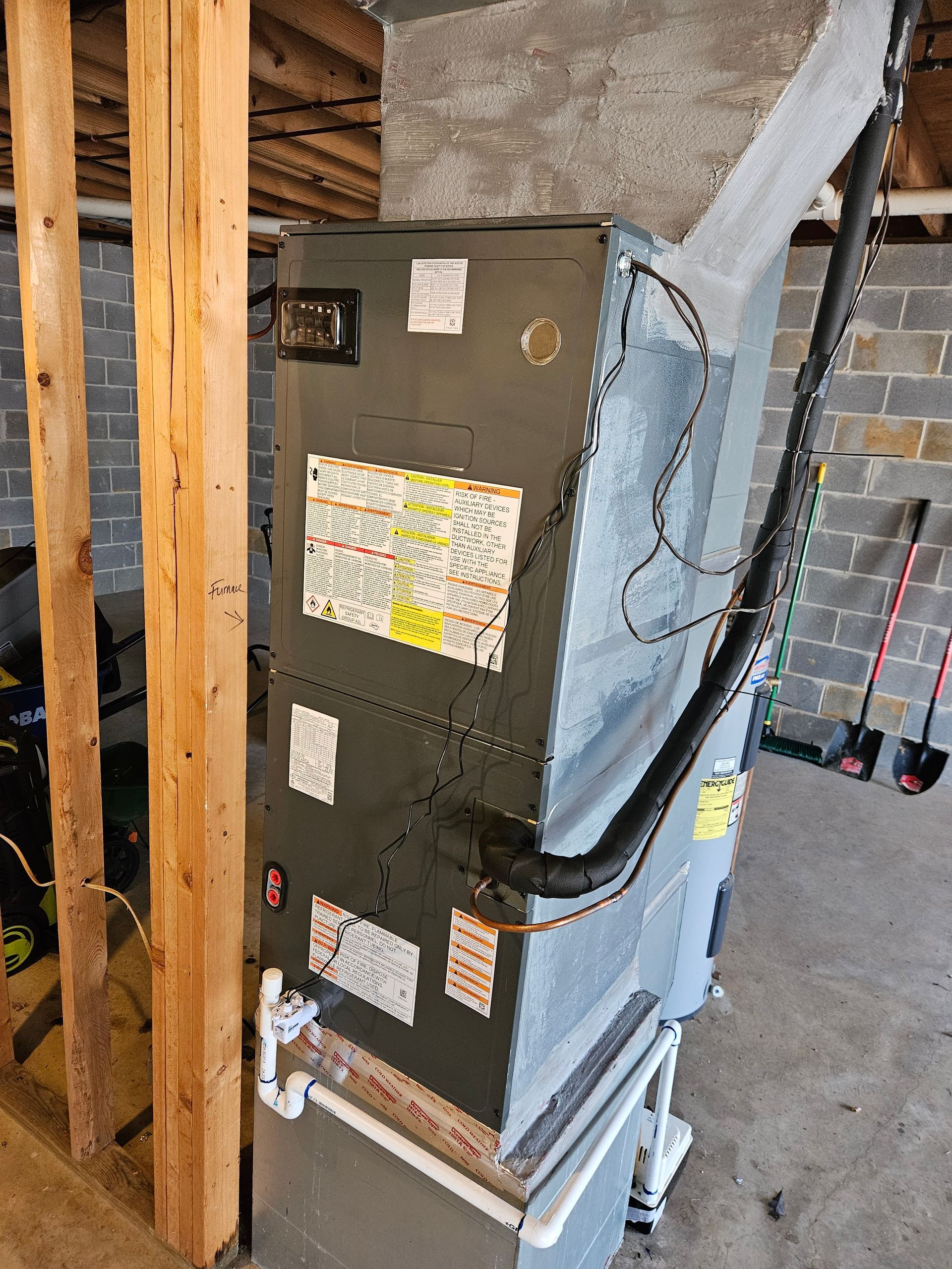 A furnace in a basement setting. Gray metal unit with various labels and connected to black tubing,