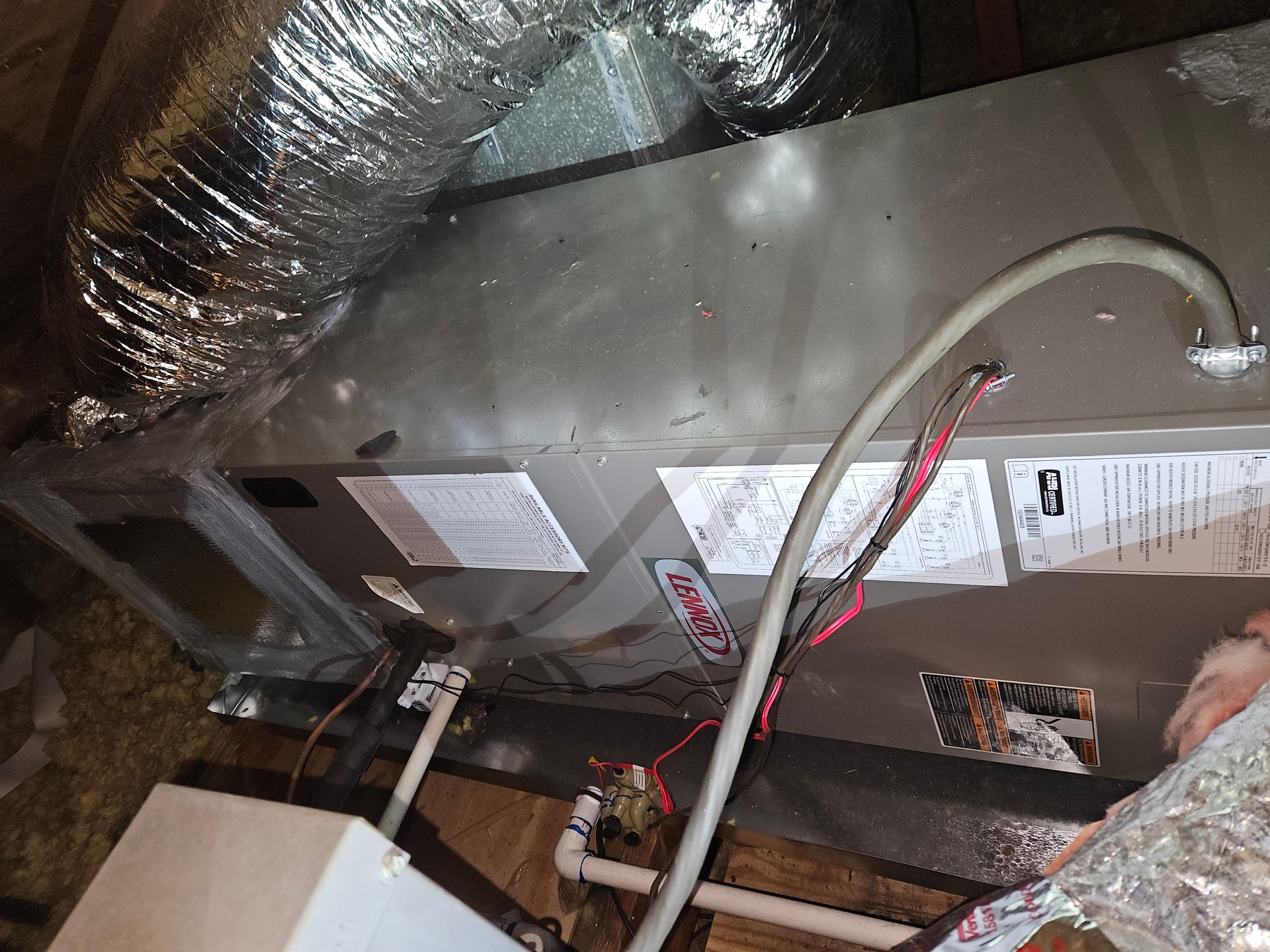 HVAC unit in an attic with ductwork, wiring, and piping visible. The unit is metallic with labels.