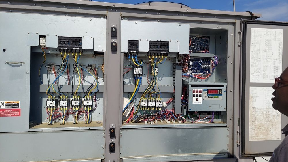 An open electrical panel with wires and components, observed by a man wearing glasses, outdoors.