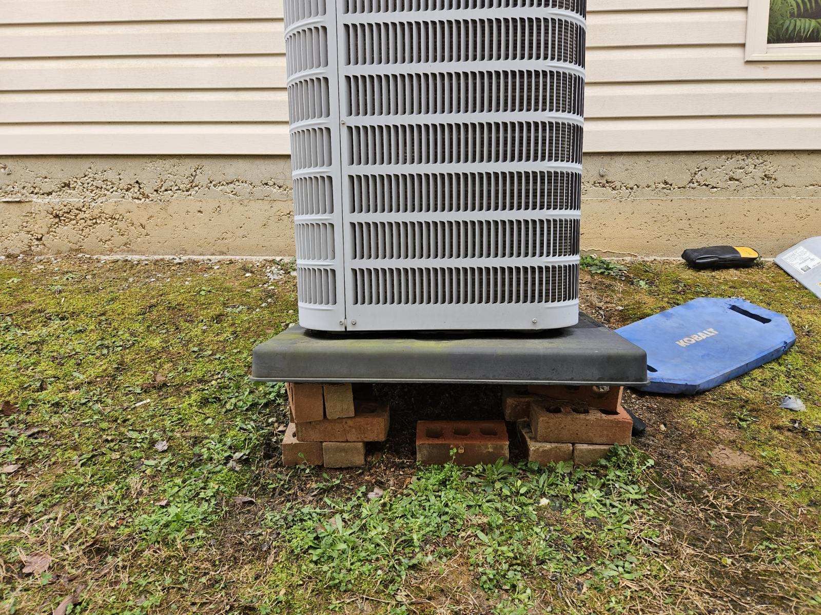AC unit resting on a black platform supported by brick. The unit sits on grass next to a house.