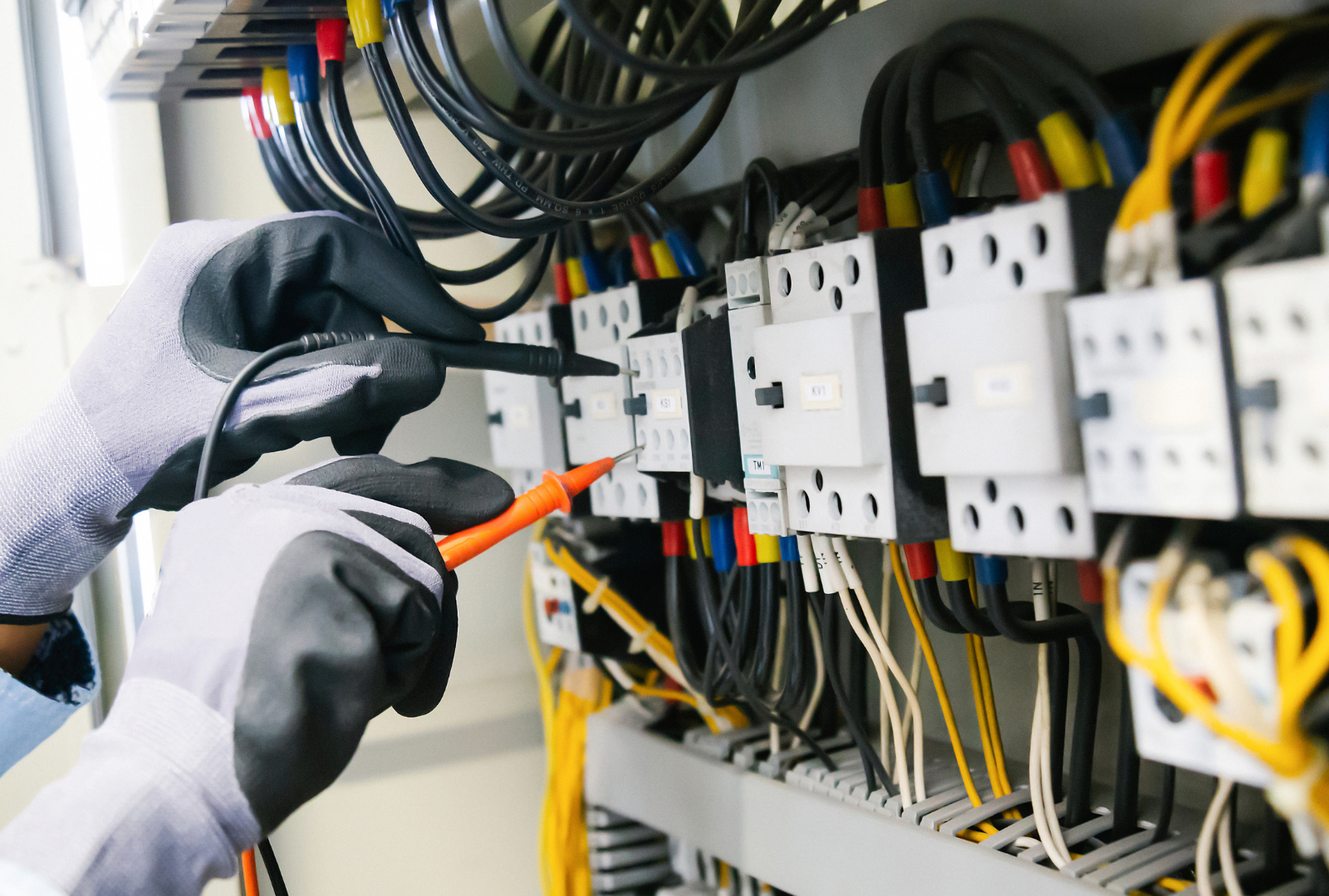 Hands in grey work gloves use multimeter probes to test connections on a row of electrical contactors in a control panel.