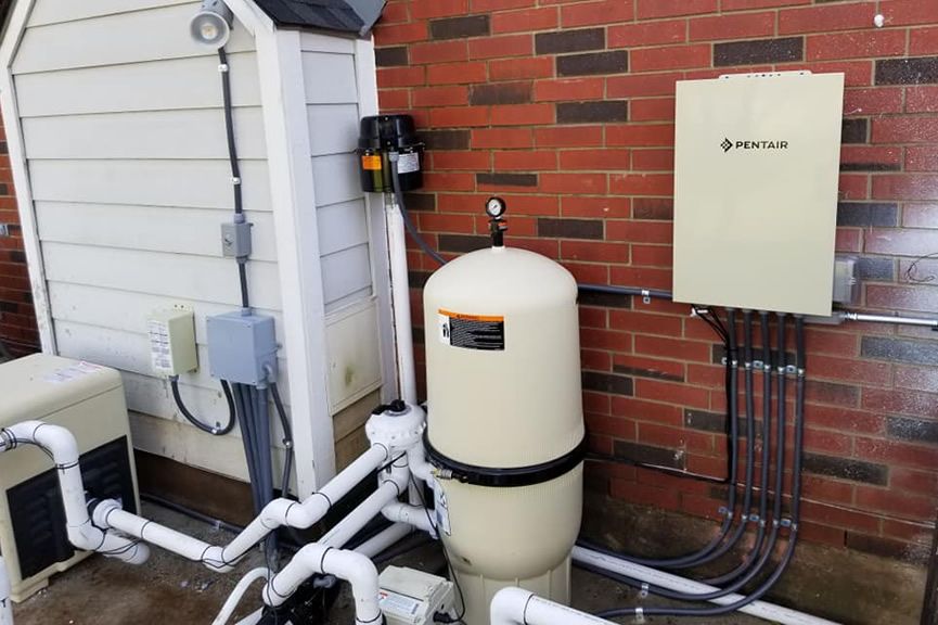 A pool filter, pump, and Pentair control box with white piping mounted against a red brick exterior wall.