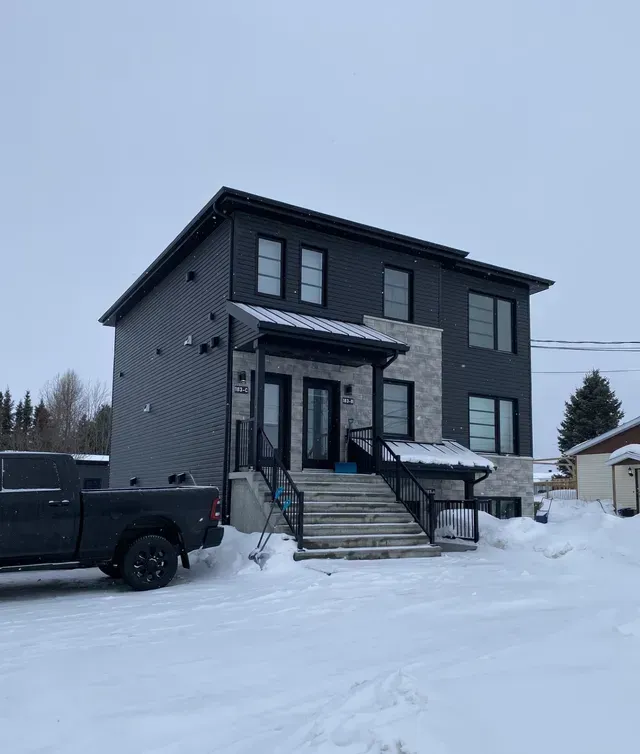 Un duplex de deux étages, couleur anthracite, avec des accents de pierre et un escalier central en béton, entouré d'une épaisse couche de neige hivernale.