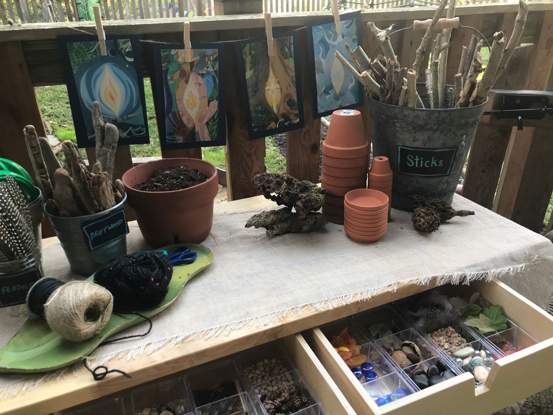 An invitation to explore and wonder... Art supplies on a wooden table, including paintings, sticks, pots, twine, and beads.