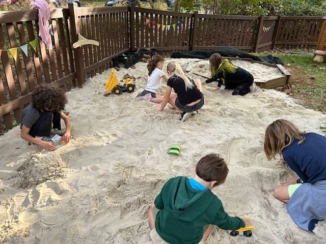 Children playing in a sandbox with toys. A wooden fence surrounds them.