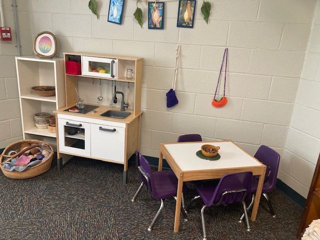 Our indoor classroom space. Play kitchen and table set in a child's playroom. White cabinets, purple chairs, art on wall.