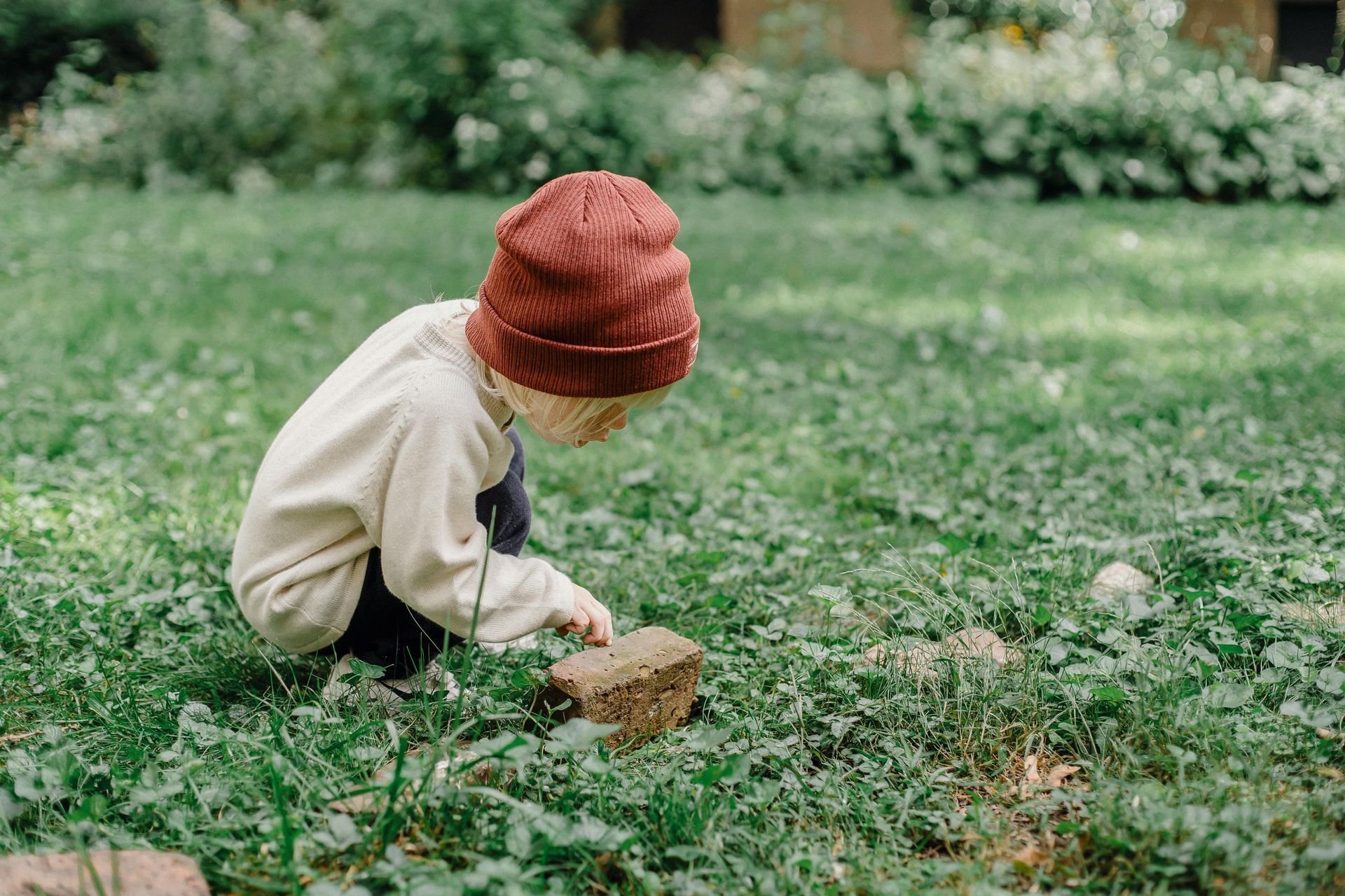 Child wearing a knit hat, examining a small brick in a grassy area outdoors.
