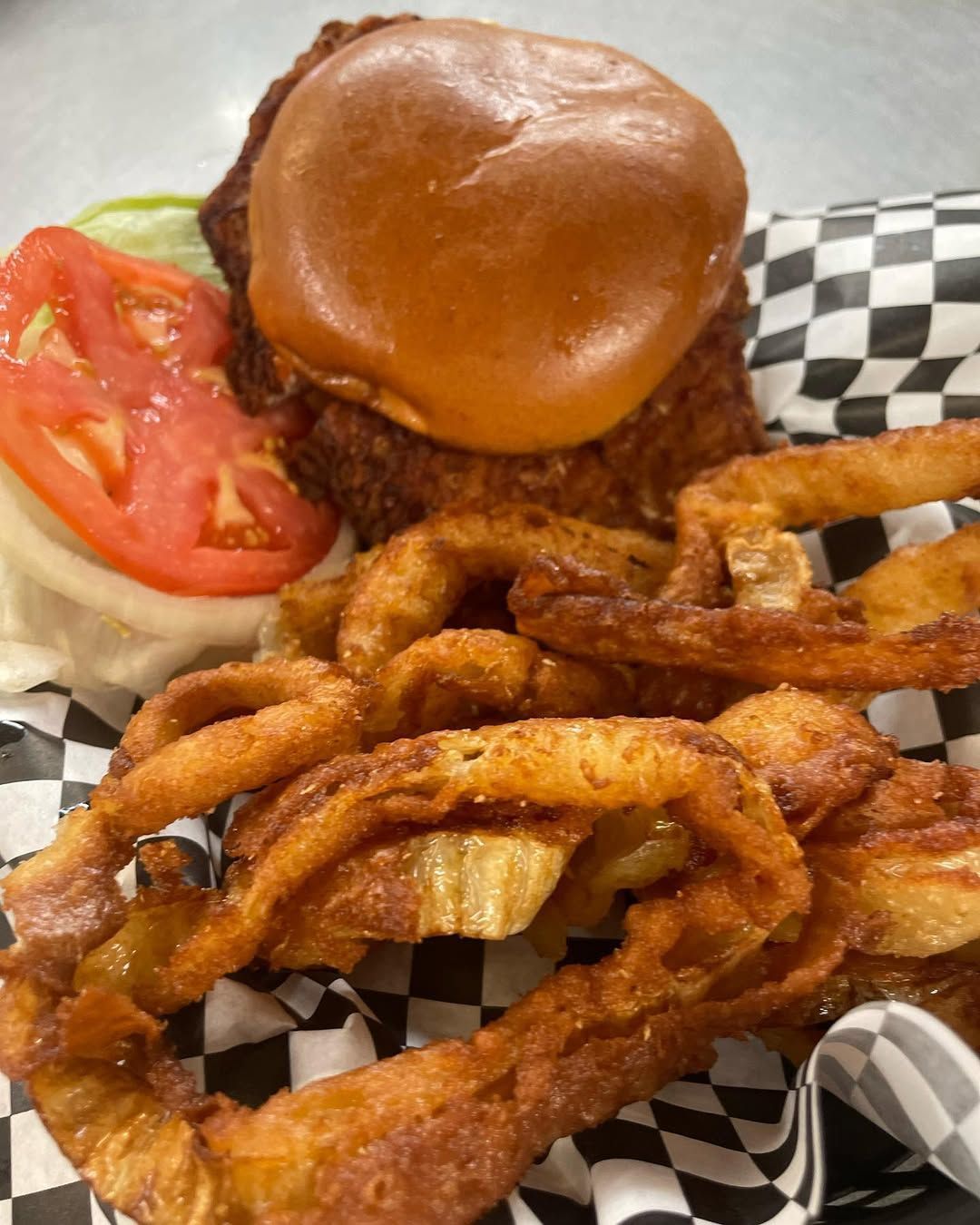 A chicken sandwich with onion rings and french fries on a checkered napkin.